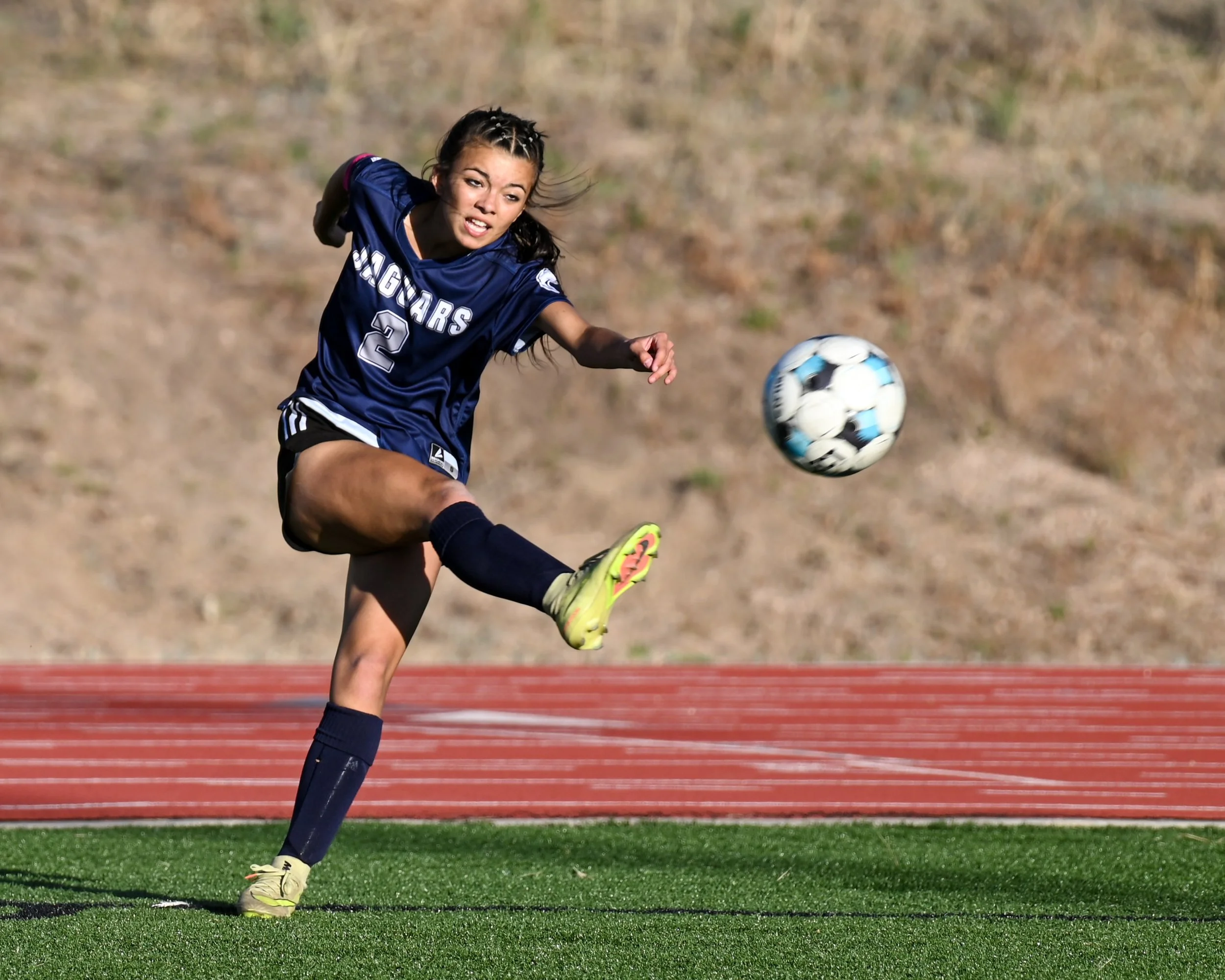 A female soccer player in a dark blue uniform with the number 2 is kicking a soccer ball on an outdoor field with a grassy background.