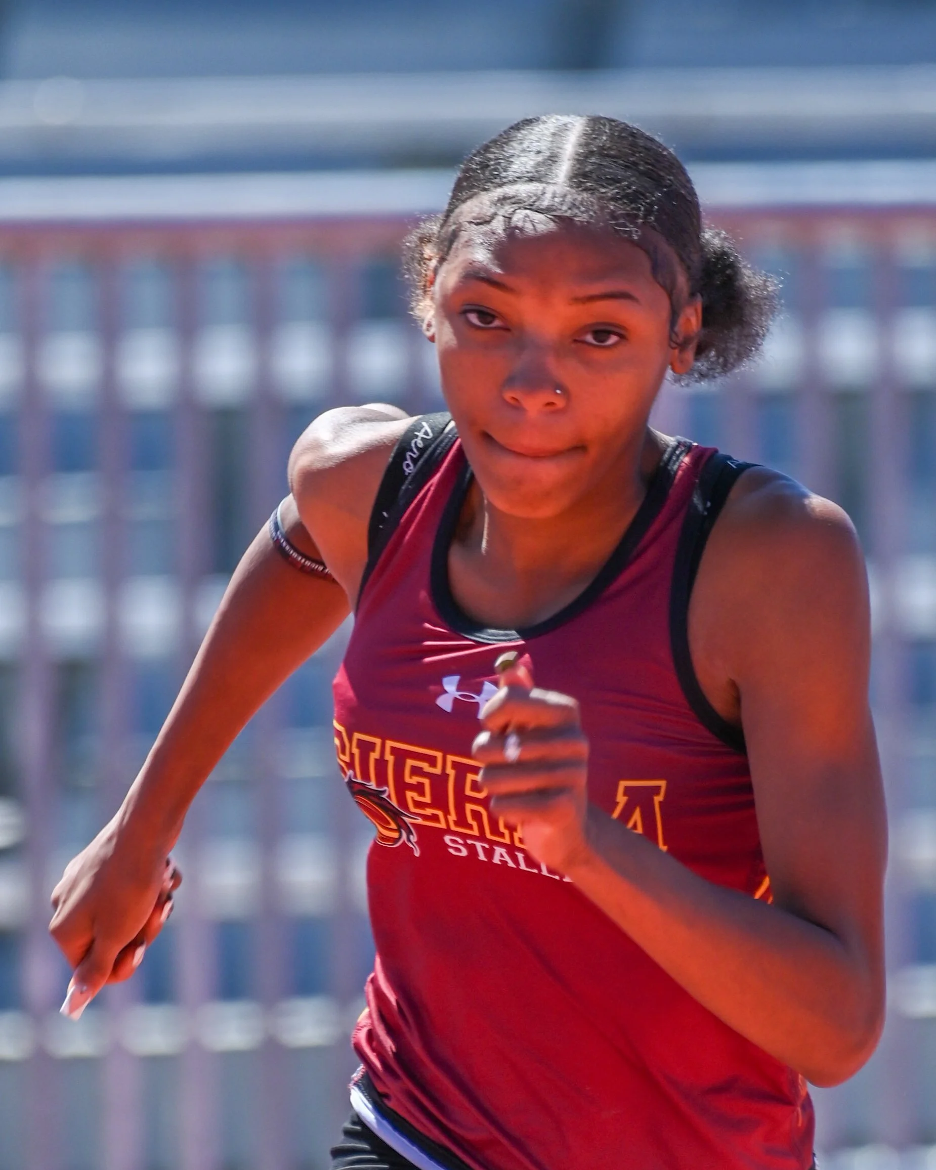 A female track athlete in a red jersey, is running outdoors in a sprinting race.