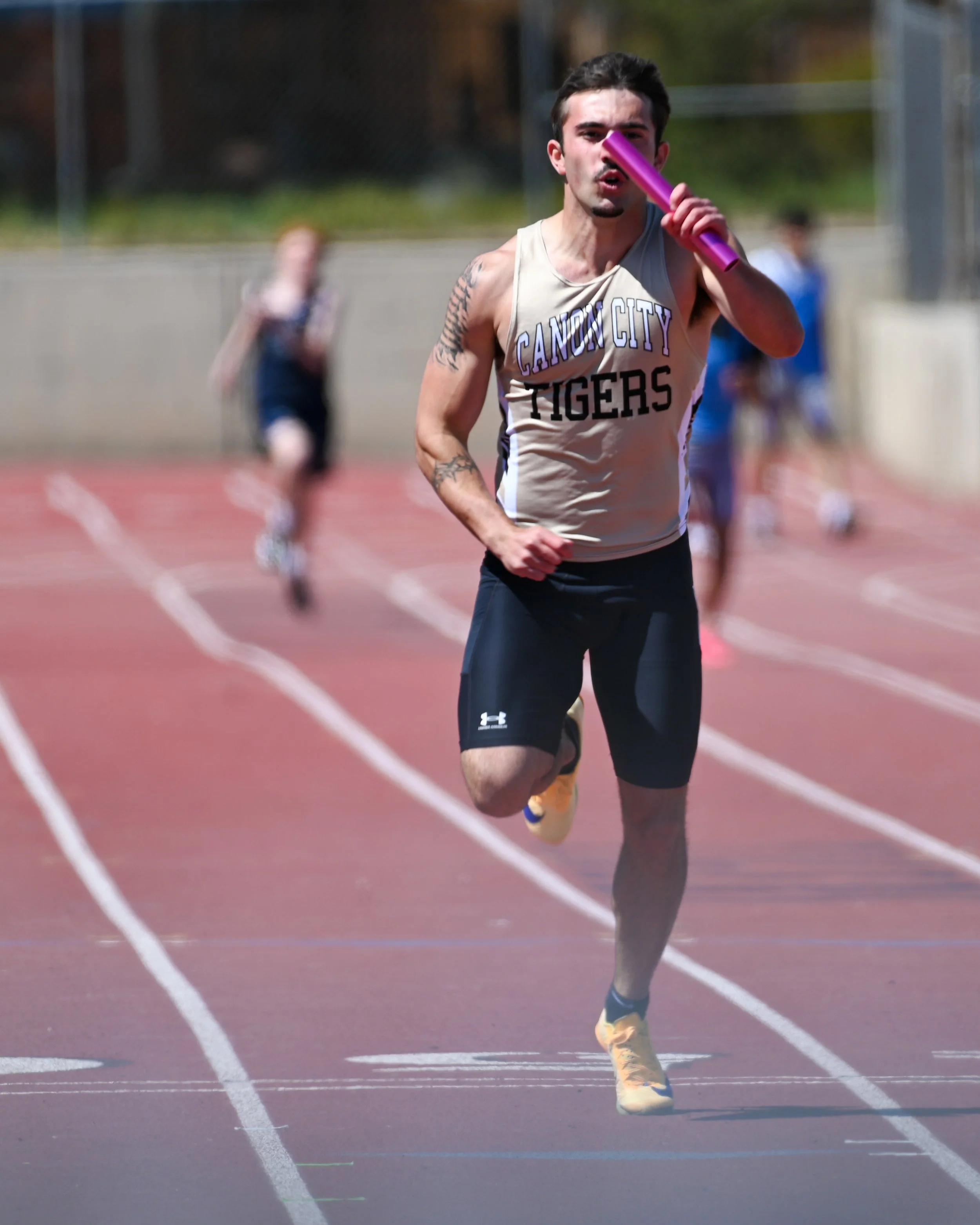 Male track athlete running on a red track, holding a purple baton, wearing a beige tank top with 'Cañon City Tigers' written on it, black shorts, and yellow running shoes with blue accents.