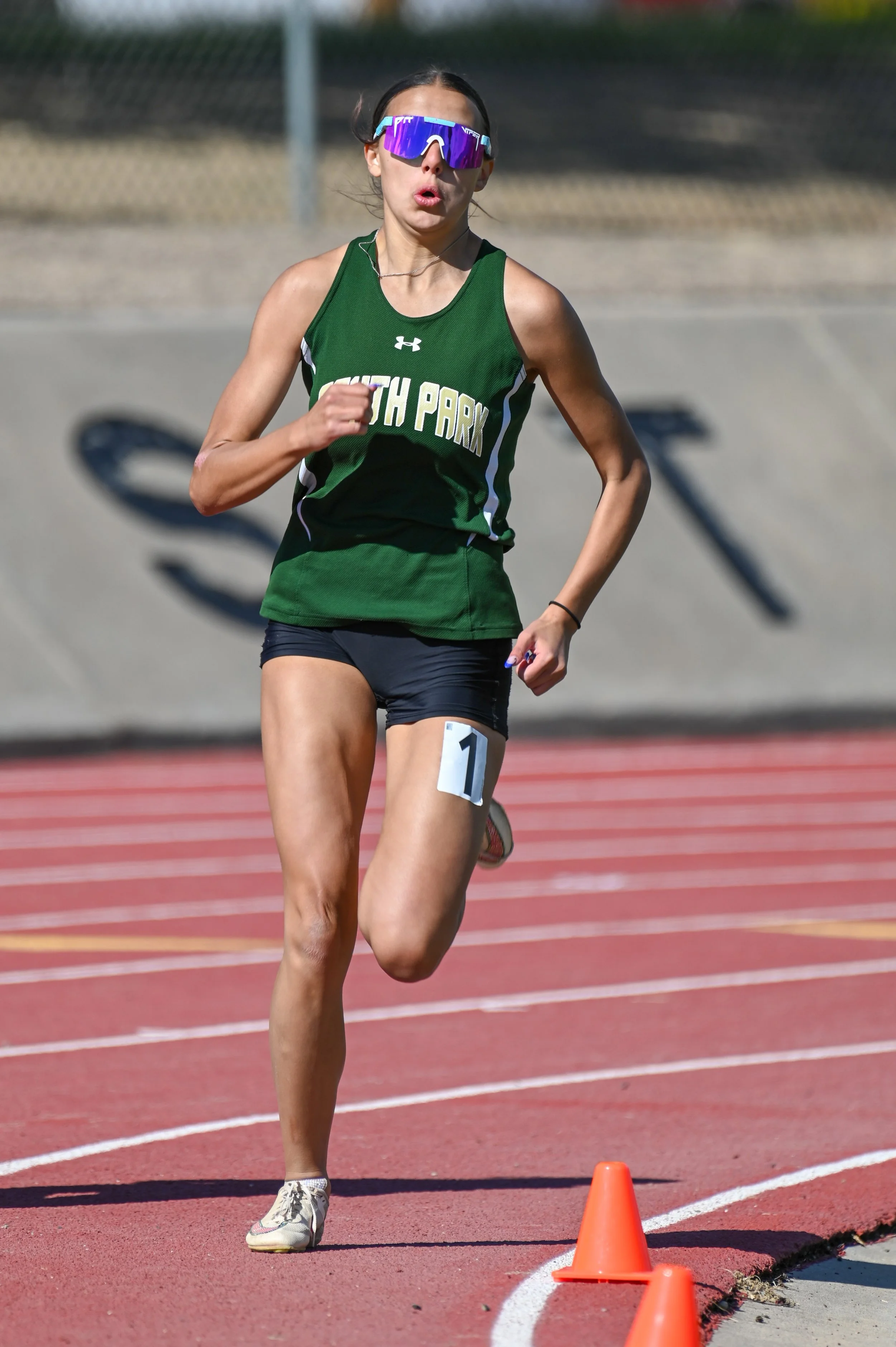 A female runner on a track during a race wearing sunglasses, a green top that says 'SOUTH PARK,' and black shorts. She is running around orange cones placed along the lane.