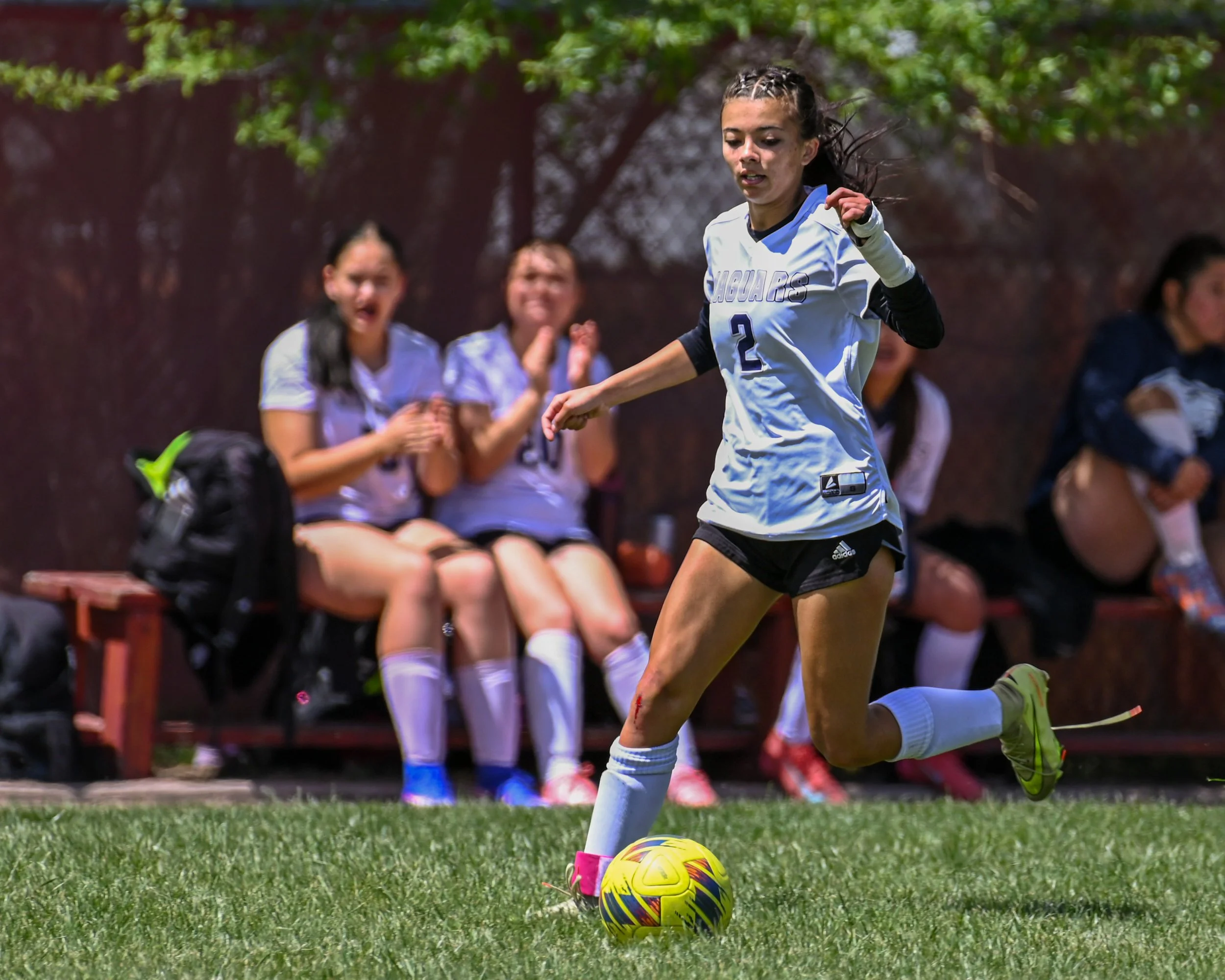 Female soccer player kicking a yellow and blue soccer ball during game, with players on the bench clapping in the background.