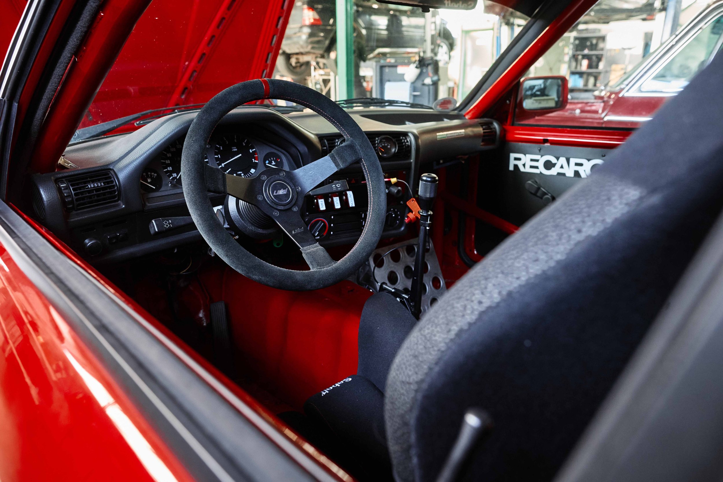 Innenansicht eines roten Rennwagens, der Rennlenkrad, Tachometer, Schalthebel, Instrumentenanzeigen und schwarze Sitze zeigt. Der Hintergrund ist eine Werkstatt mit Autos und Werkzeugen.