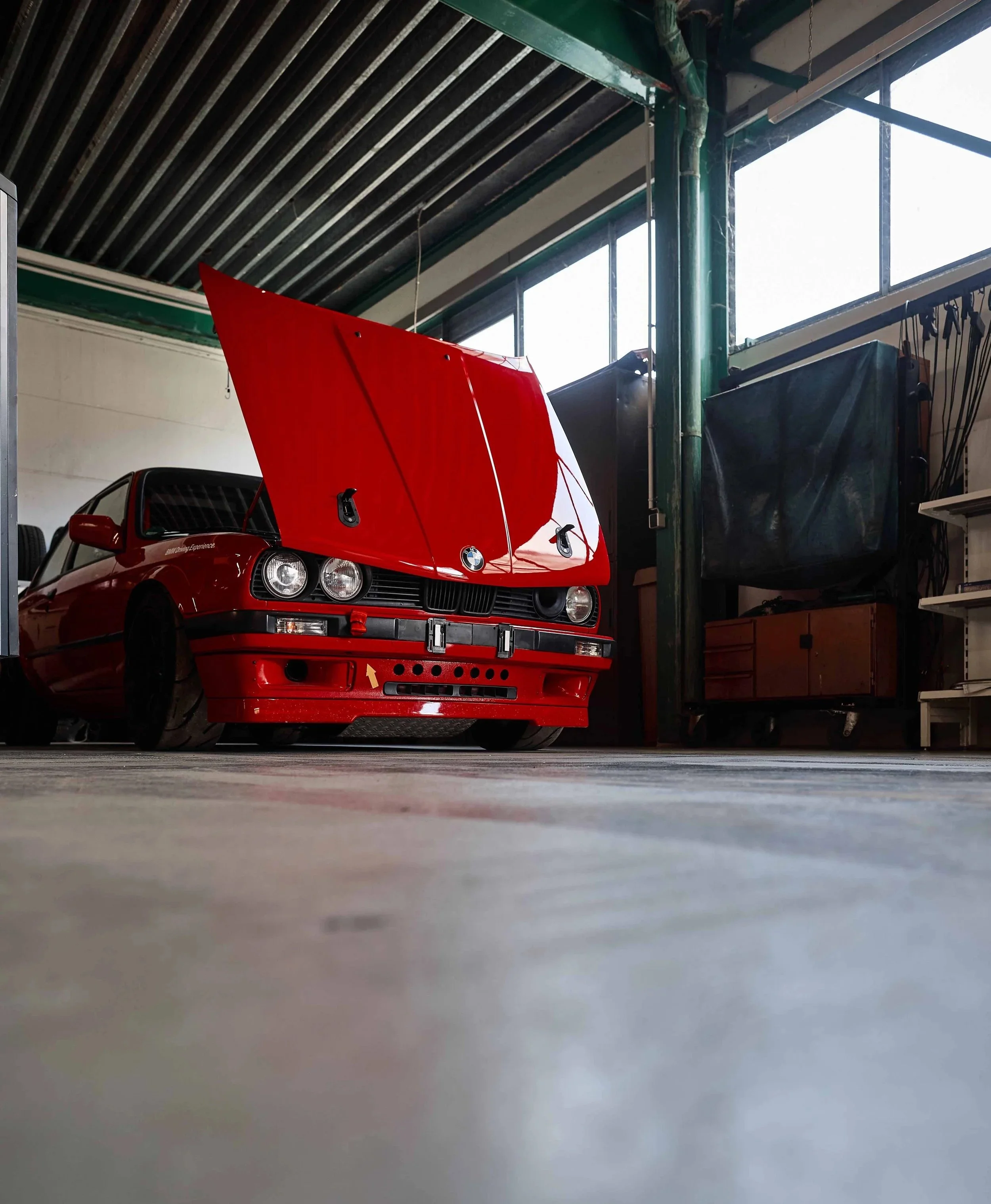 Ein rotes Auto mit offenem Motorhaube in einer Werkstatt oder Garage, von einem niedrigen Blickwinkel aufgenommen.
