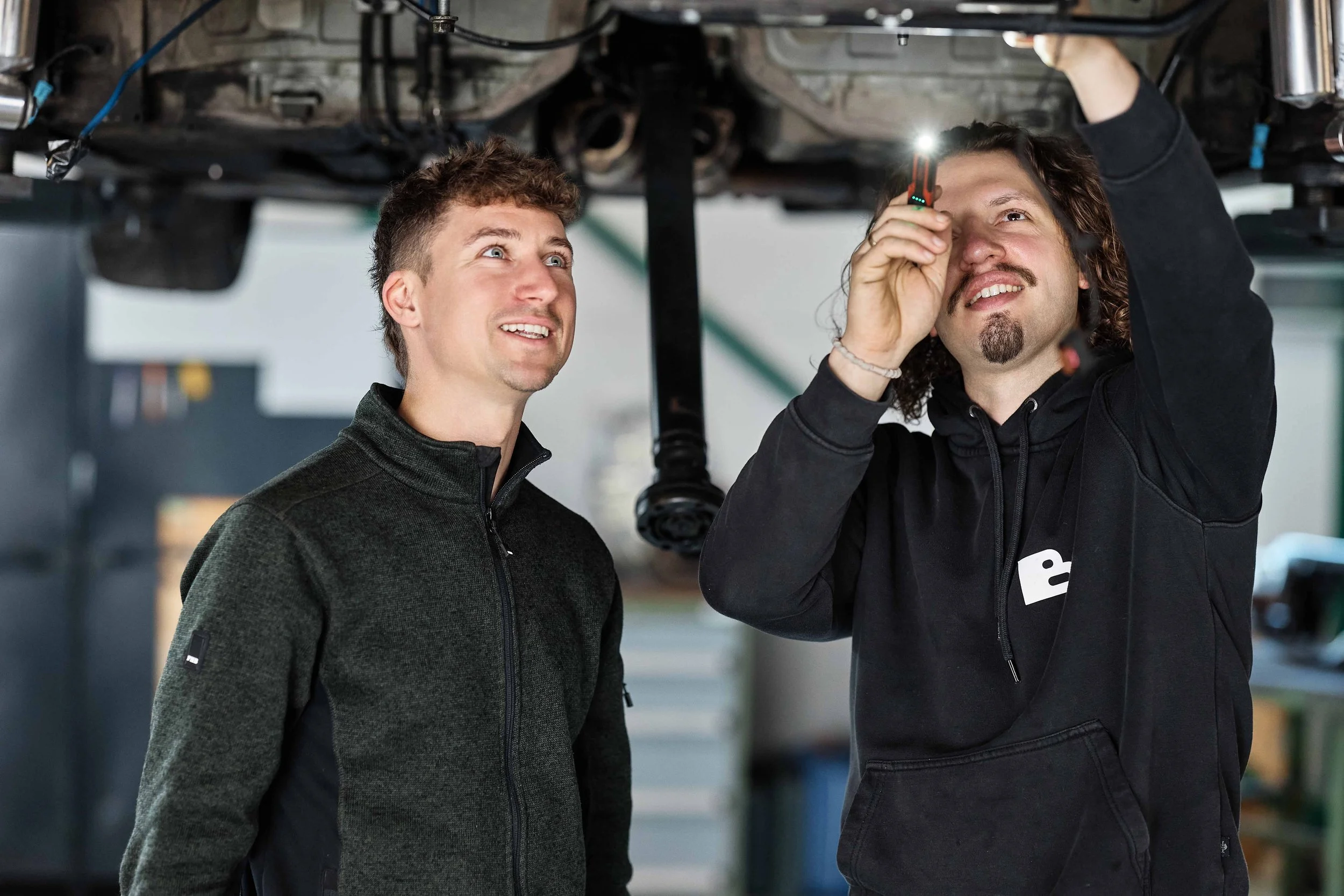 Zwei Männer arbeiten unter einem Auto, einer benutzt ein Messgerät, der andere schaut zu, in einer Werkstatt.