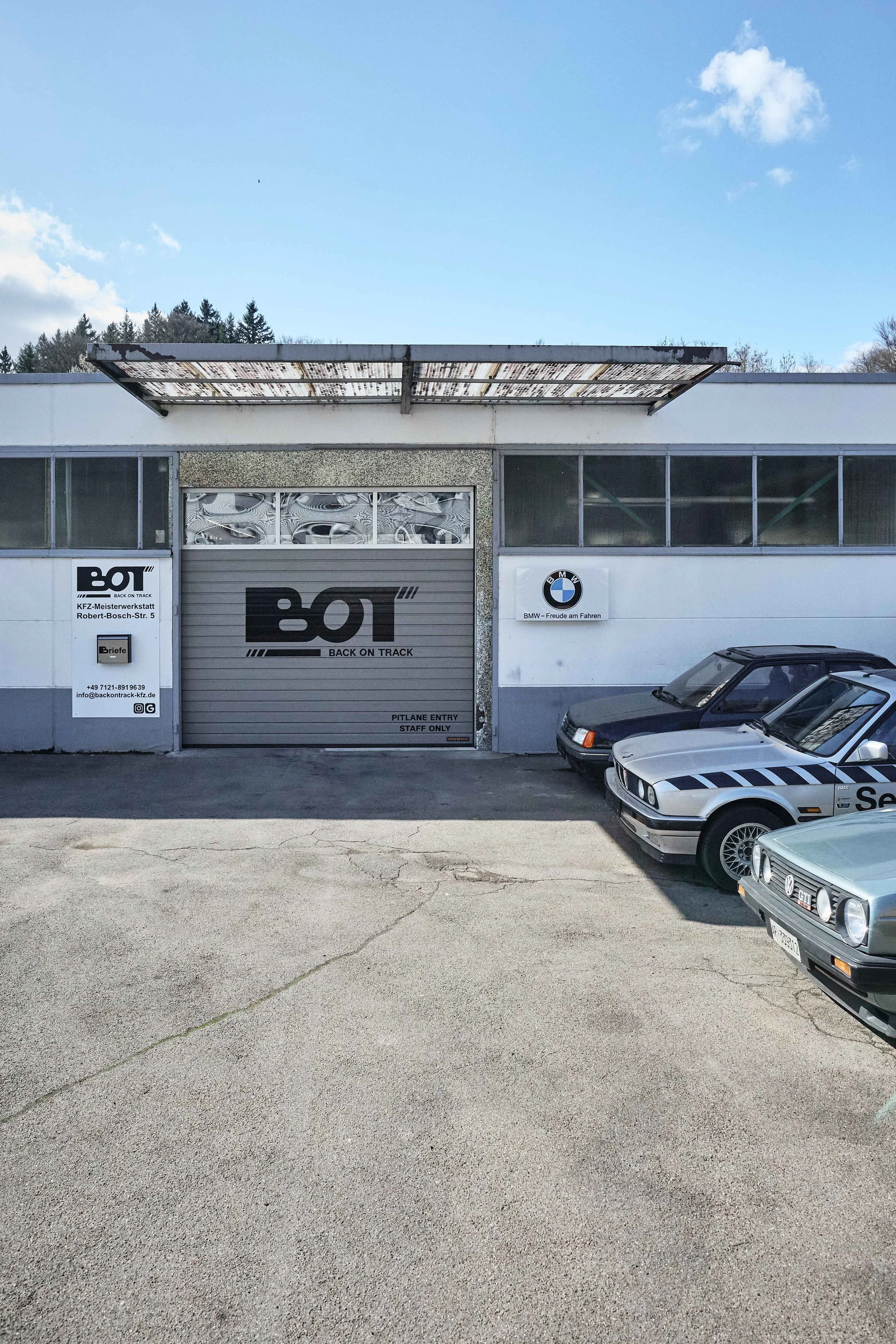 Außenansicht eines Auto-Servicelagers mit mehreren geparkten klassischen Autos, darunter BMW und Volkswagen, in der Nähe eines Gebäudes mit Firmenlogos.
