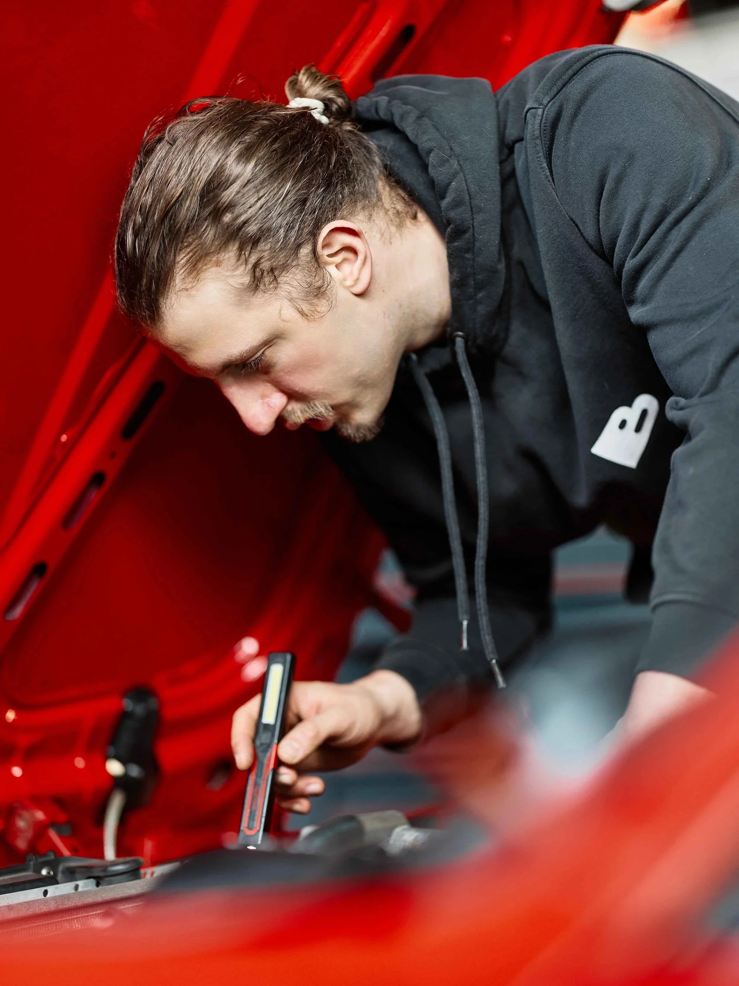 Ein Mann arbeitet an einem roten Fahrzeug, möglicherweise ein Auto, und benutzt ein Werkzeug, um an einer Stelle im Motorraum zu arbeiten.