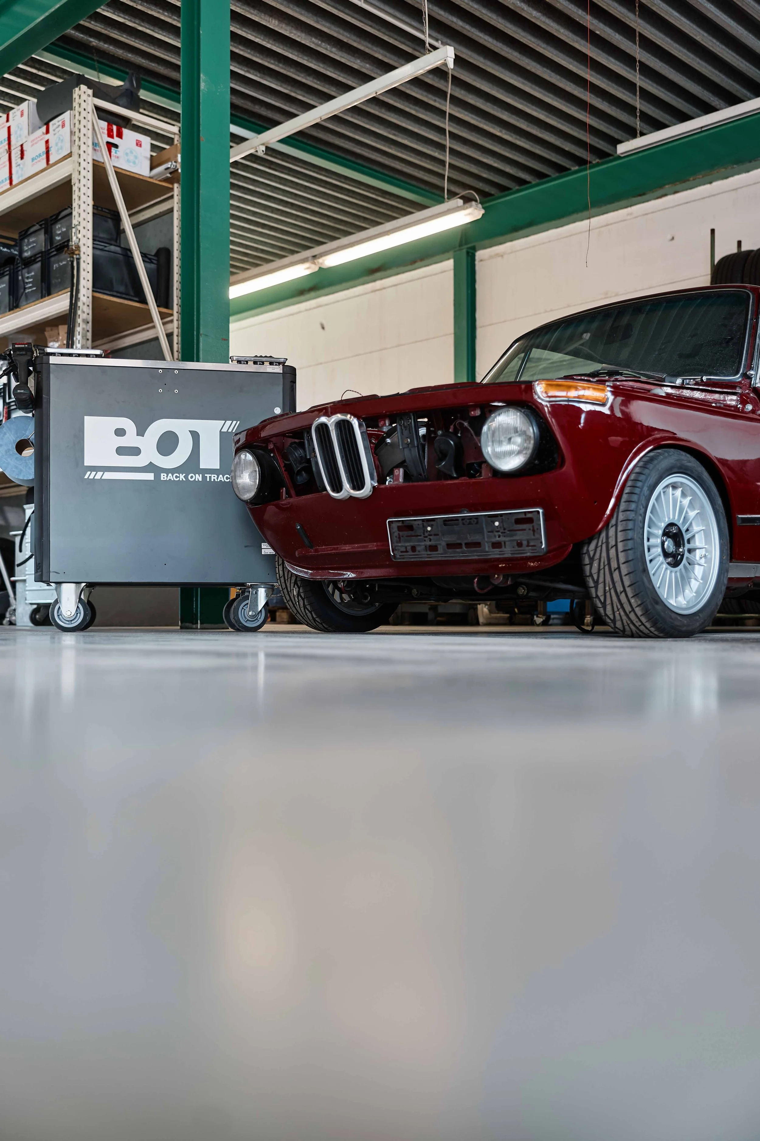 In einem Lagerraum steht ein rotes Auto, das repariert wird, neben einem Werkzeugwagen mit dem Logo 'BO.T'.