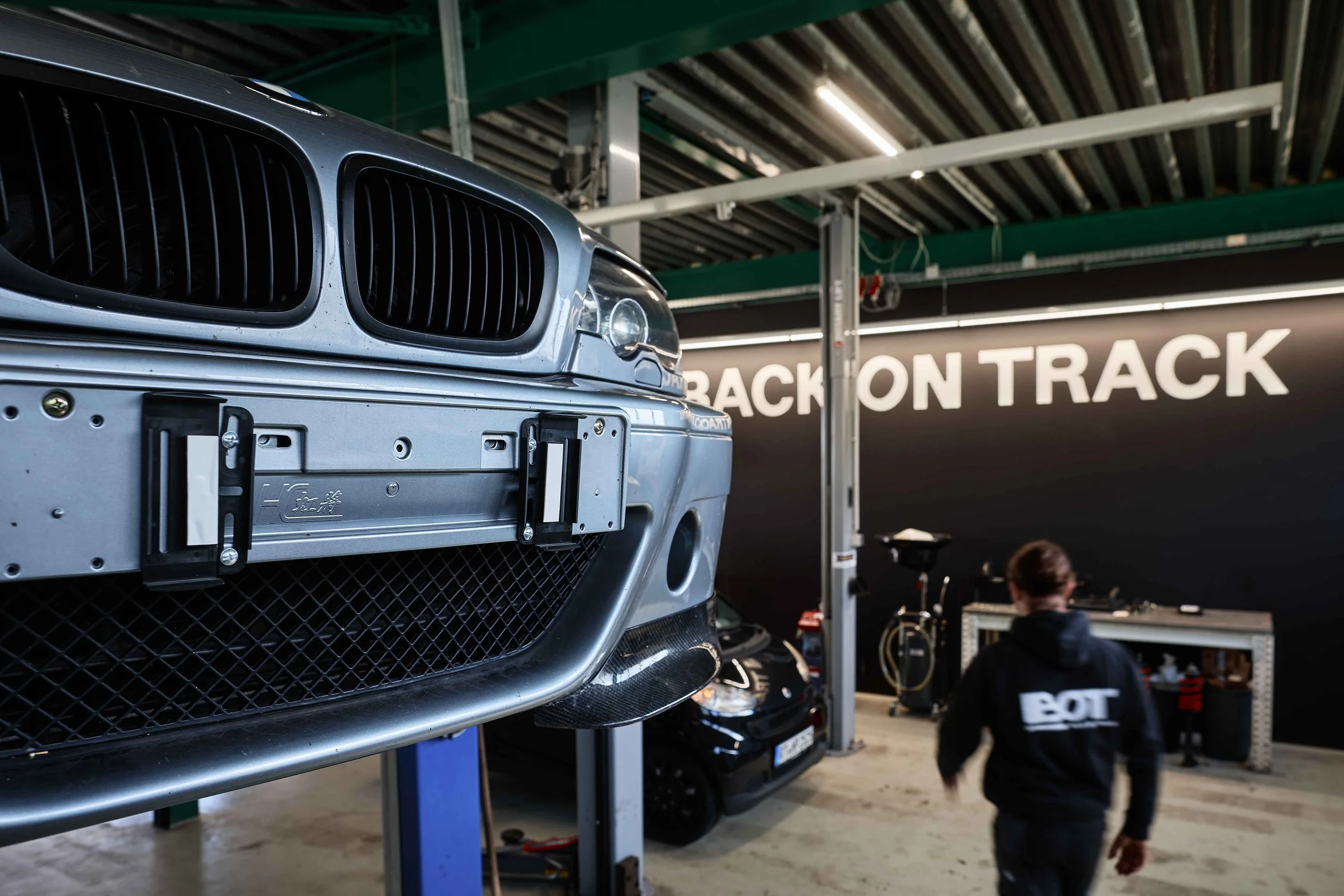 Vorderansicht eines Autokühlergrills an einem silbernen Fahrzeug, das in einer Werkstatt auf einem Kran montiert wird. Im Hintergrund sind Werkstattteile, ein schwarzer Banner mit weißen Buchstaben und eine Person mit einem schwarzen T-Shirt mit weißen Buchstaben, die nach unten schaut.