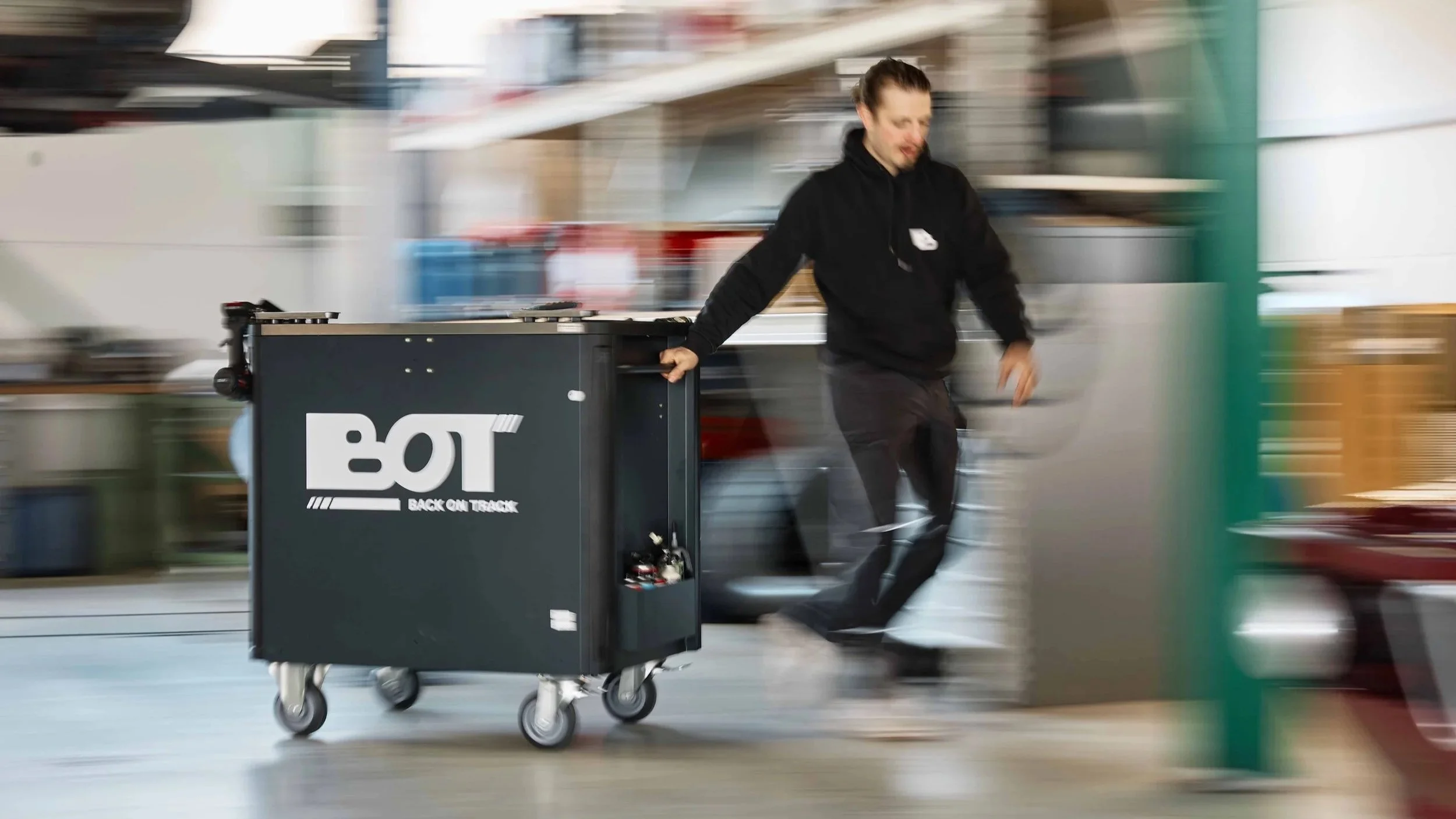 Mann schiebt einen schwarzen Gepäckwagen mit dem Logo 'BOT' im Hintergrund eines Lagerhauses oder Bahnhofs.