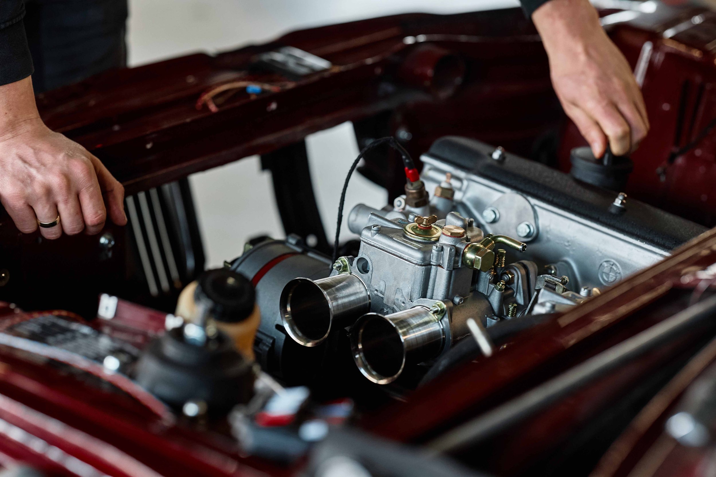 Nahaufnahme eines Automotormotors in einem roten Motorraum. Zwei Hände, eine mit Ehering, arbeiten am Motor.
