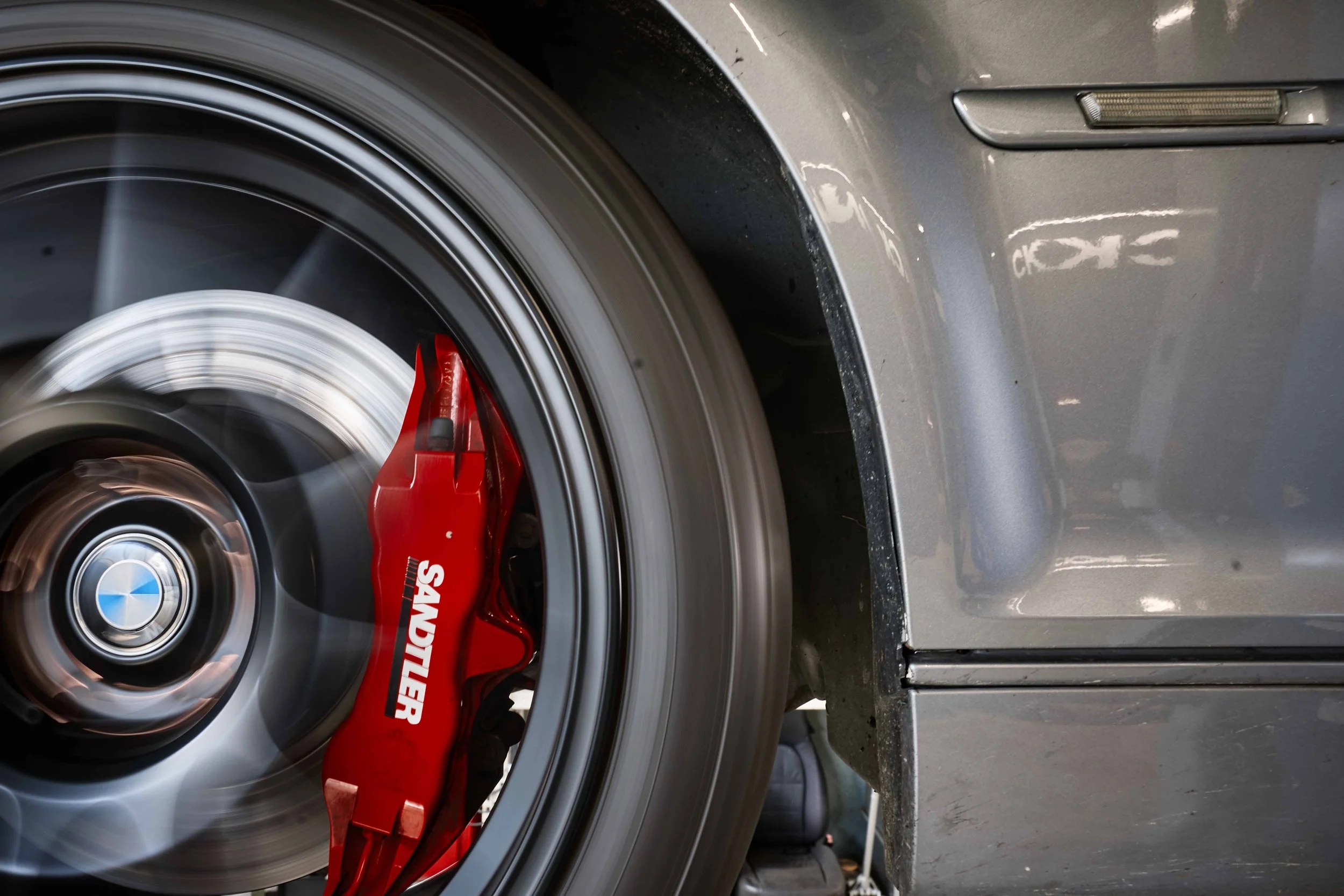 Nahaufnahme eines Autoreifens mit laufendem Rad, roter Bremssattel mit weißen Aufdruck und grauem Autolack im Hintergrund.