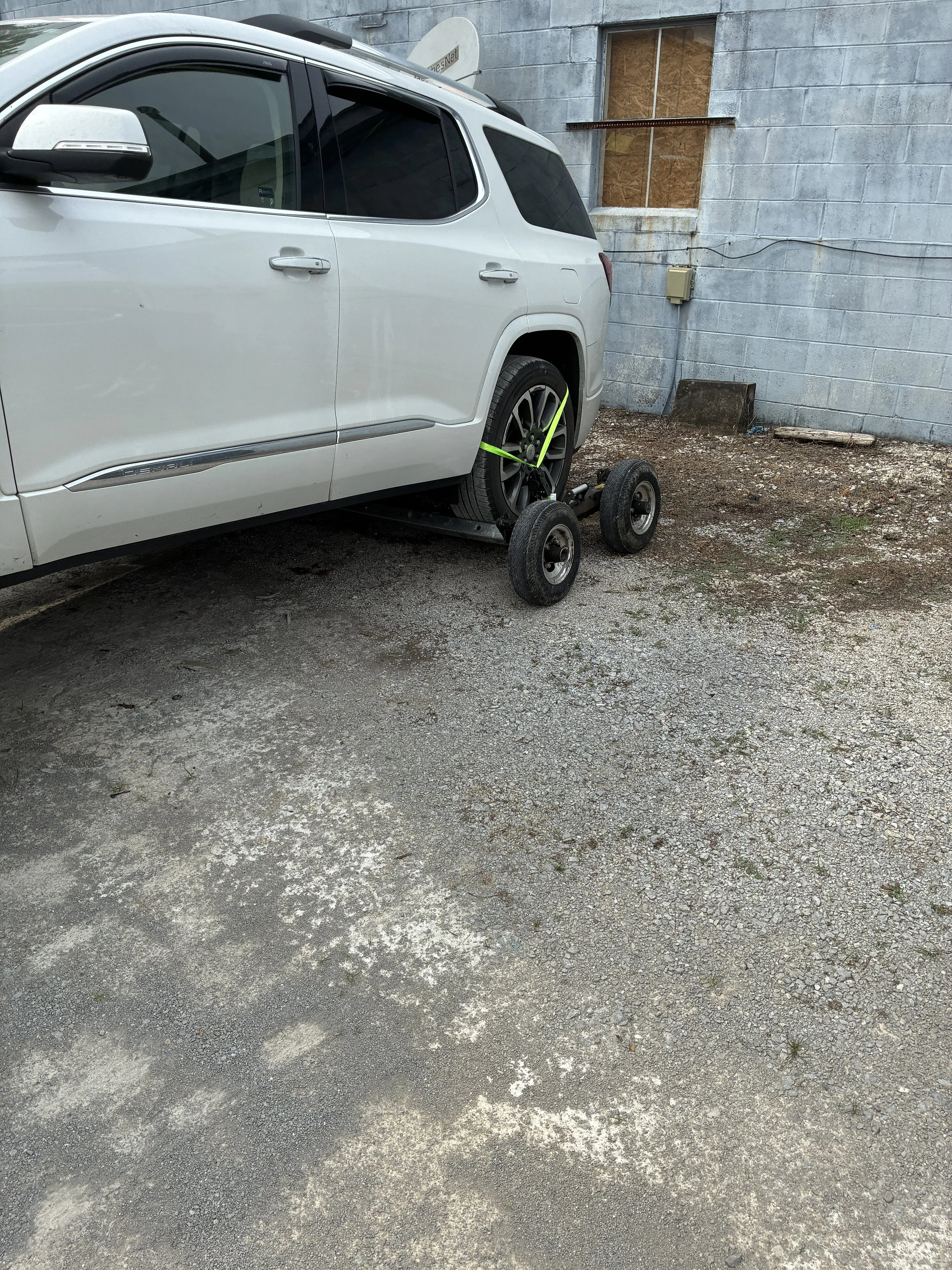 A white SUV with a black rim wheel missing, secured with a green strap, parked outside a gray cinder block building with a boarded-up window and electrical box on the wall.