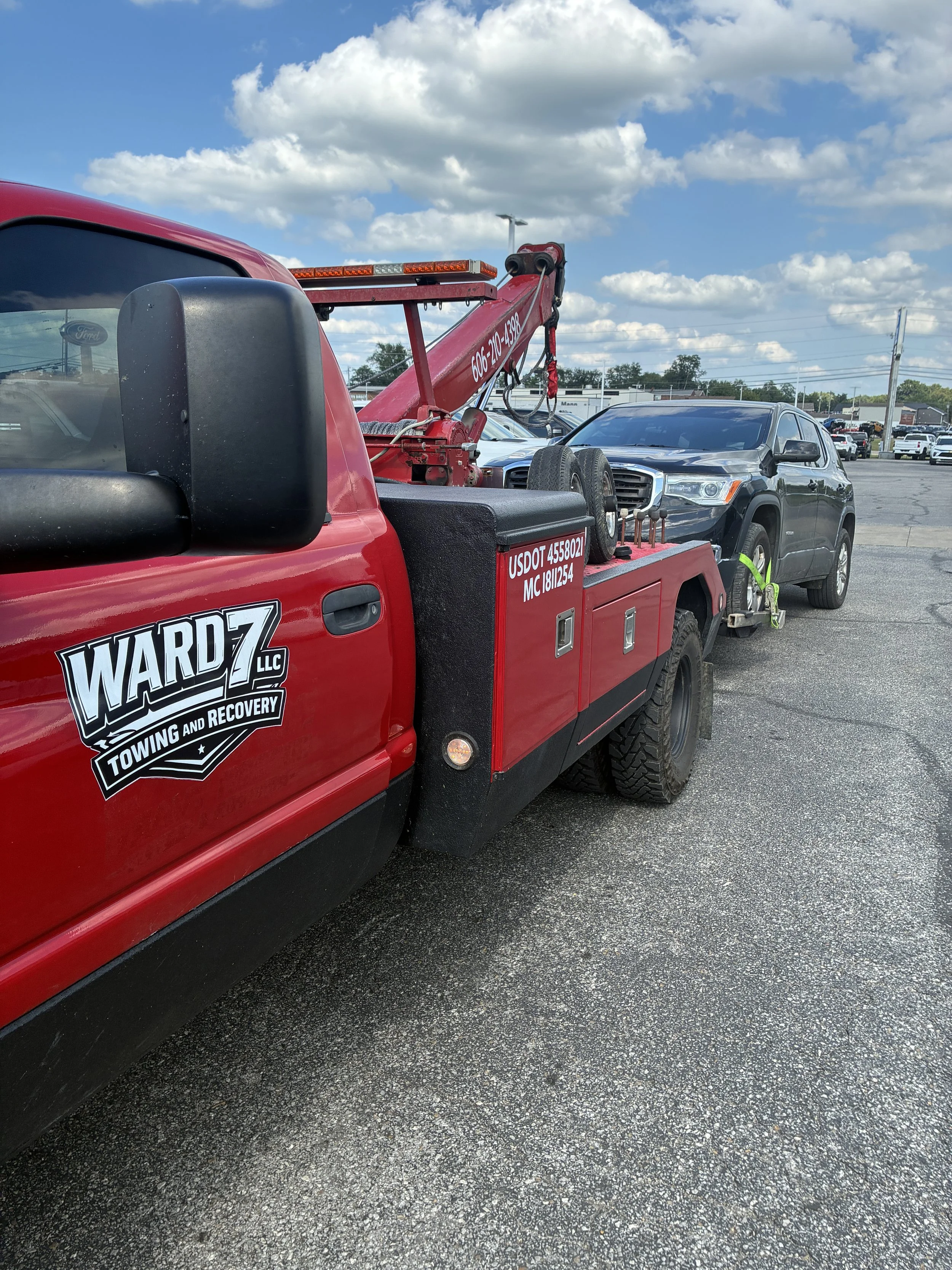 A red tow truck with WARD7 LLC Towing and Recovery logo parked in a lot with a black SUV attached on flatbed. The scene features a cloudy sky and other cars in the background.