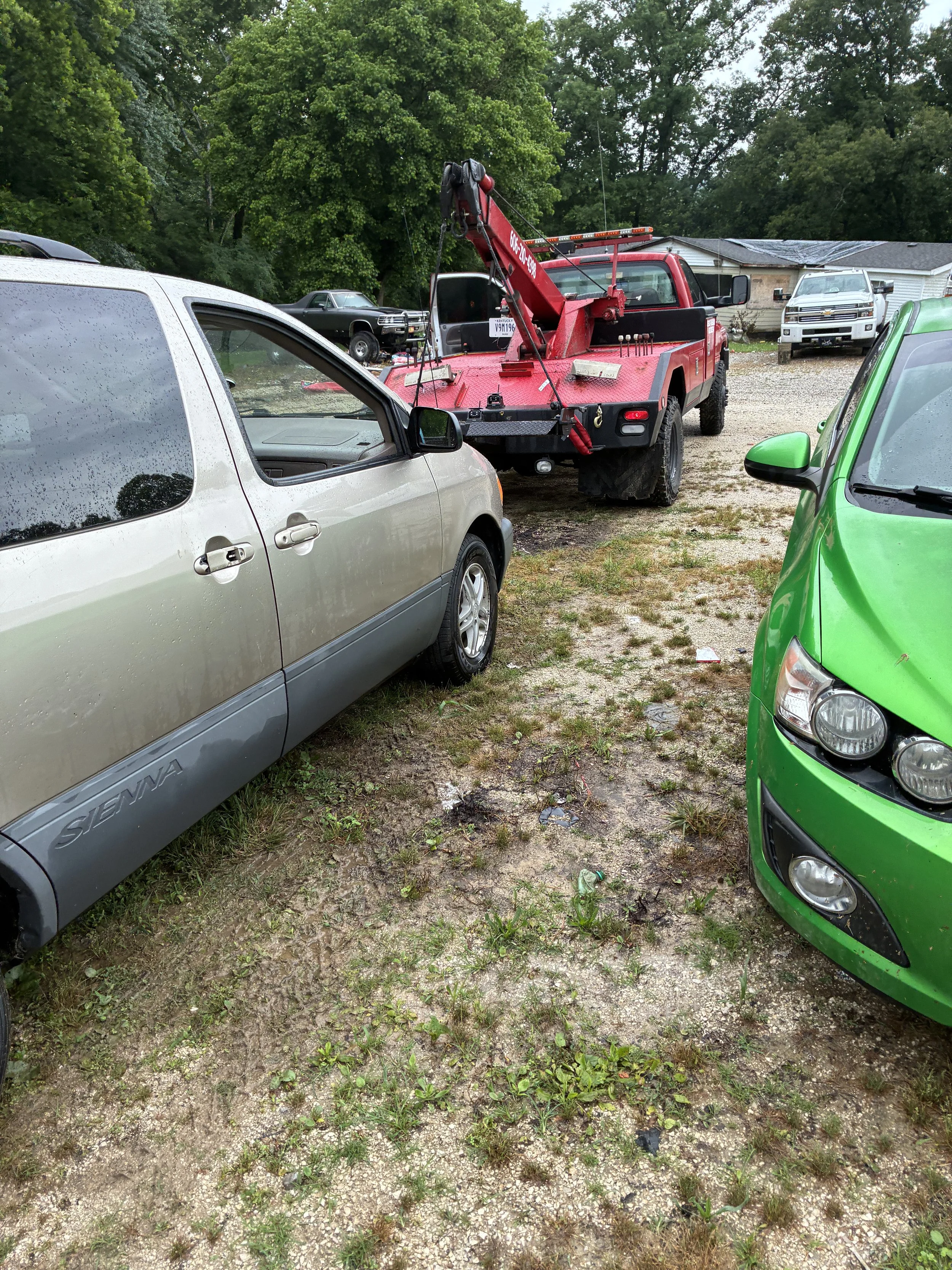 A red tow truck parked on a dirt lot with several other vehicles around, including a silver minivan in the foreground, a green car on the right, and a white pickup truck in the background, with trees and a house behind.