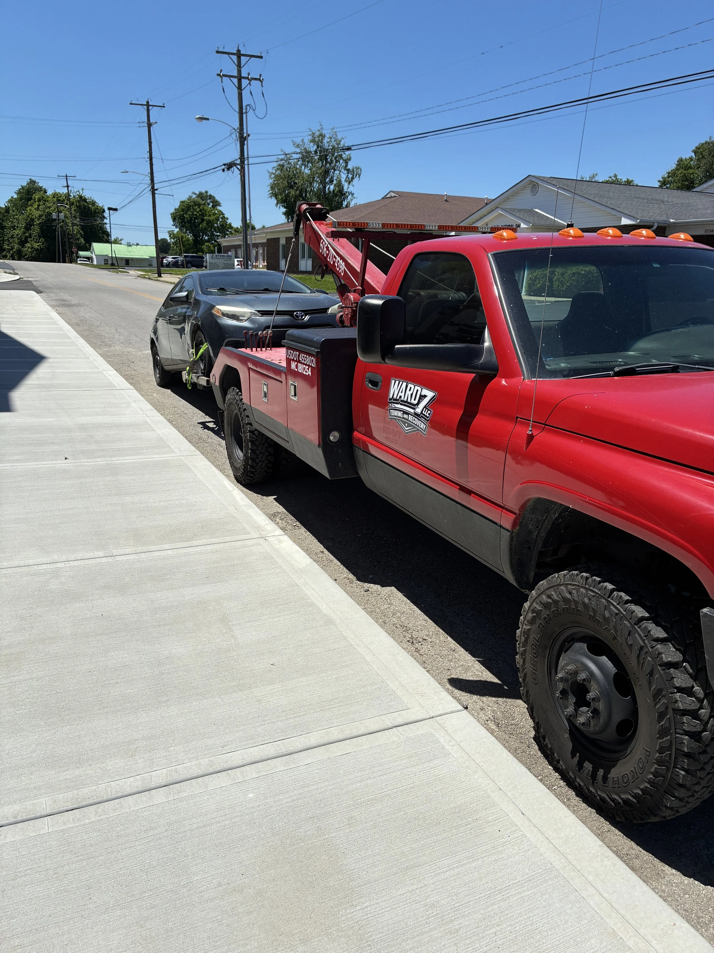 A red tow truck parked on the street with a gray car attached, in a neighborhood under a clear blue sky.