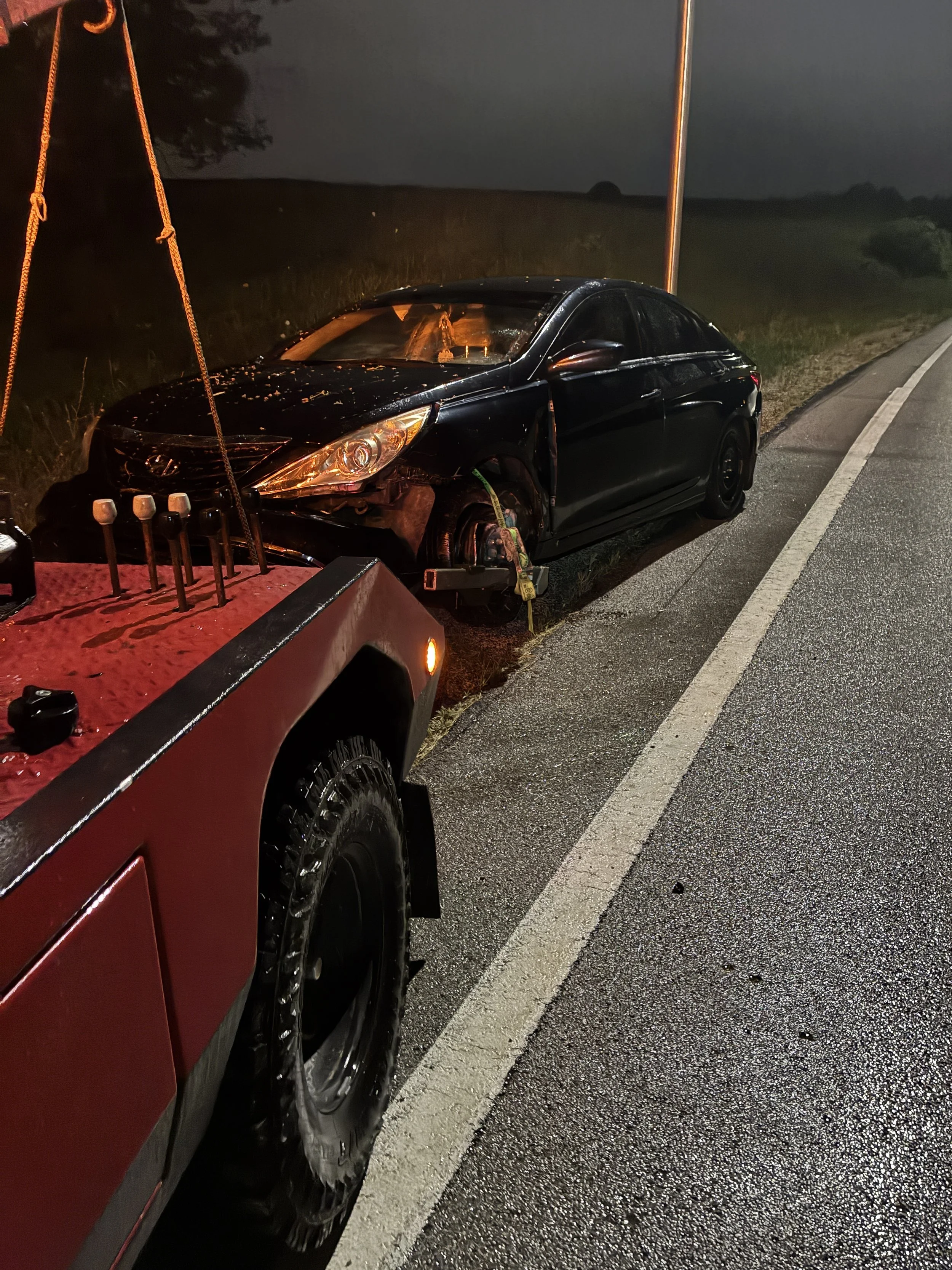 Damaged black sedan vehicle on the side of a dark, wet road at night, with a tow truck nearby.