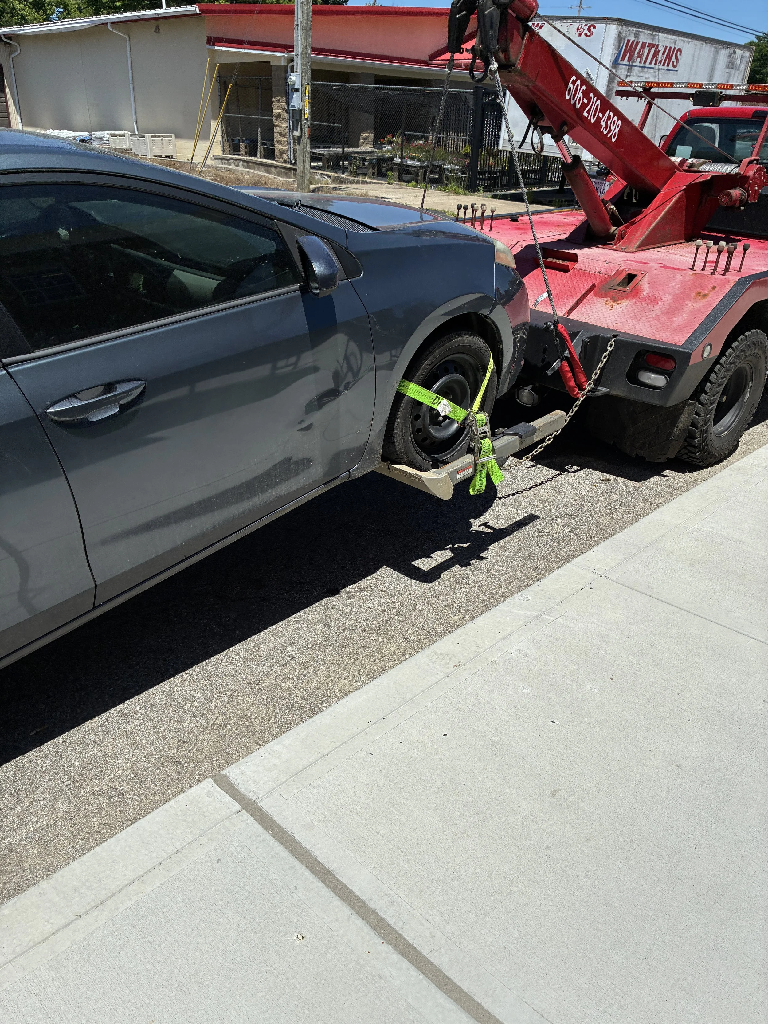 A car parked on the side of the street is being towed by a red tow truck. The tow truck has a large boom arm and is secured to the car's front wheel with a green strap and chains. The car is gray and the tow truck is red.