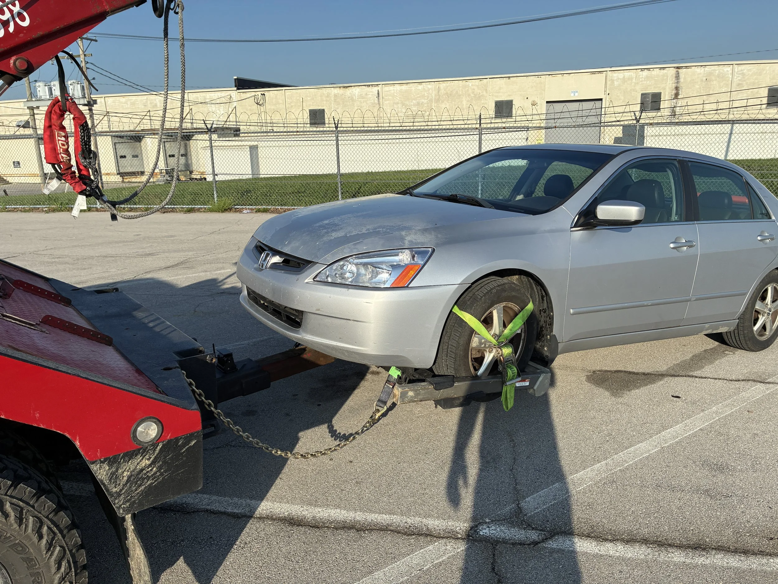 A silver Honda sedan is being loaded onto a tow truck with a red body, using green straps to secure the car. The background shows a beige building with doors, windows, barbed wire fencing, and power lines under a clear blue sky.