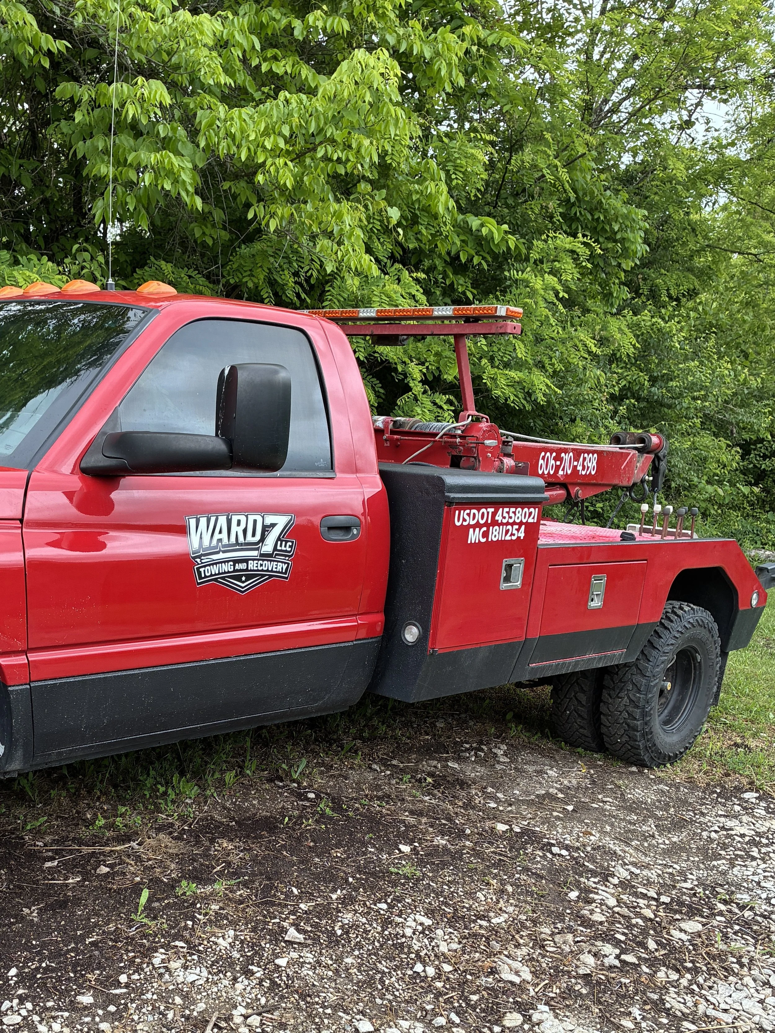 Red tow truck parked on a dirt and gravel surface with green trees in the background.