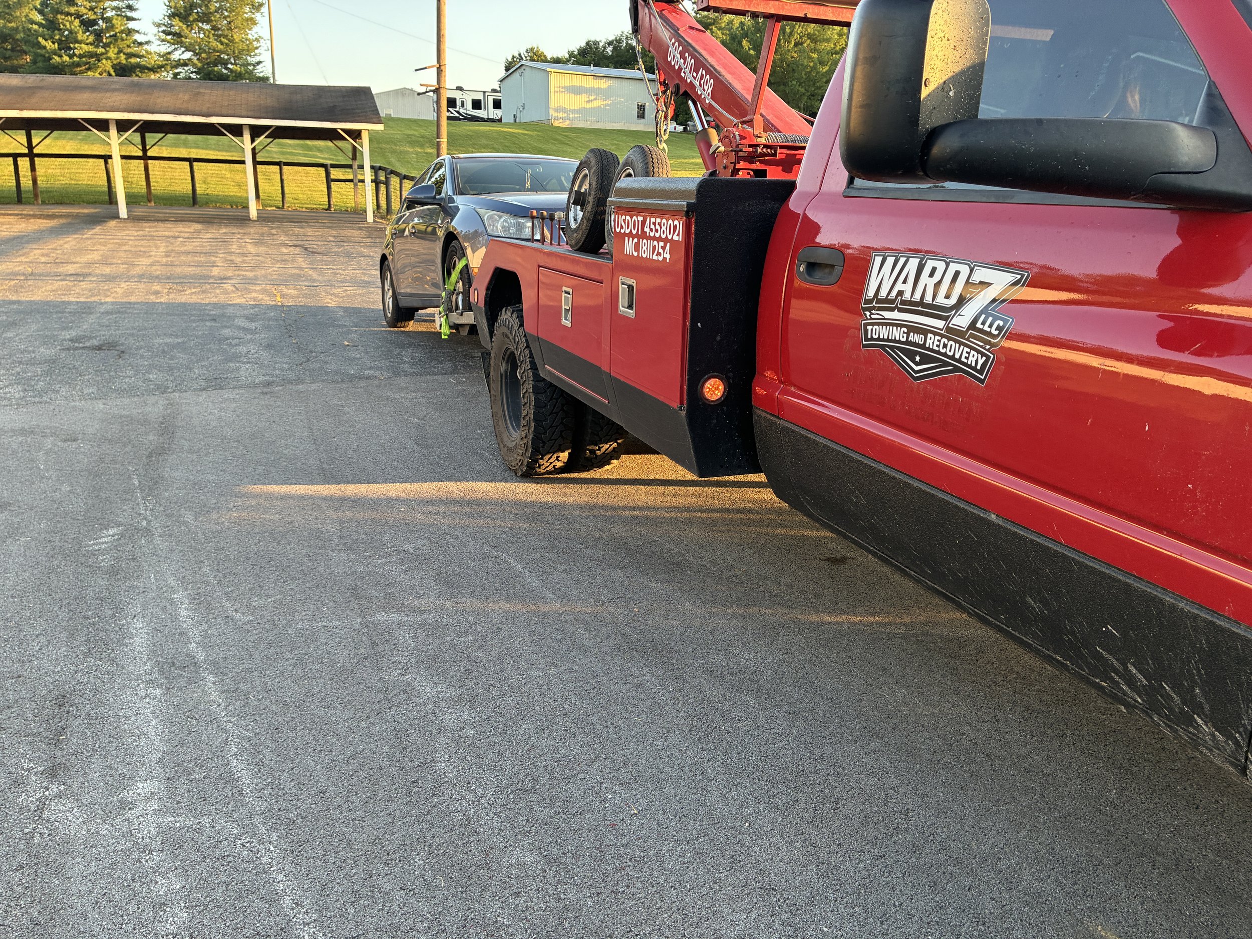 Red tow truck parked on an asphalt lot, with a gray car attached to its towing mechanism. The scene is outdoors during sunset, with green trees and a hill in the background.
