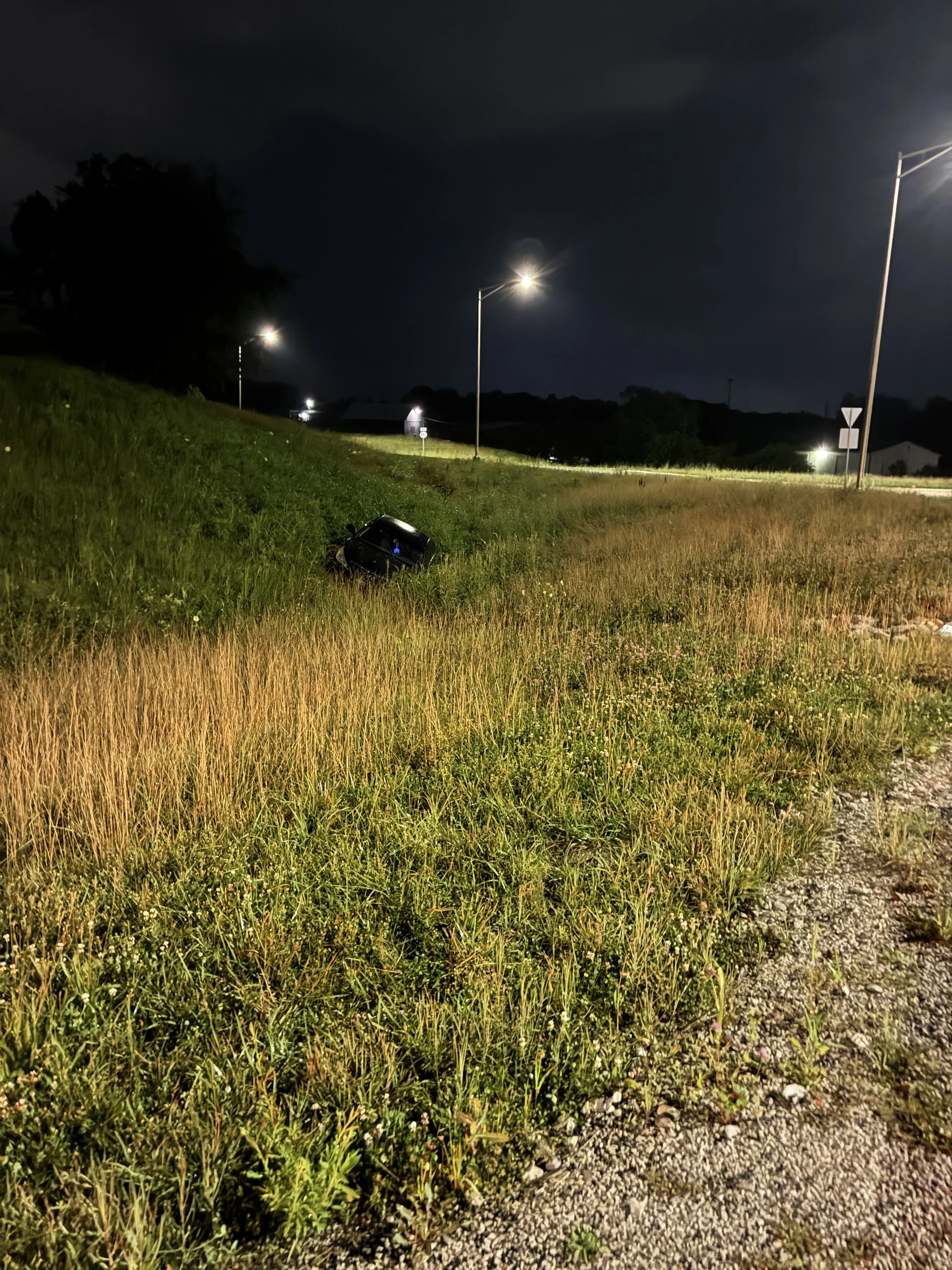 Nighttime scene of a grassy roadside area with streetlights illuminating the grass and a car partially visible on the ground.  The car failed to successfully navigate a round about.