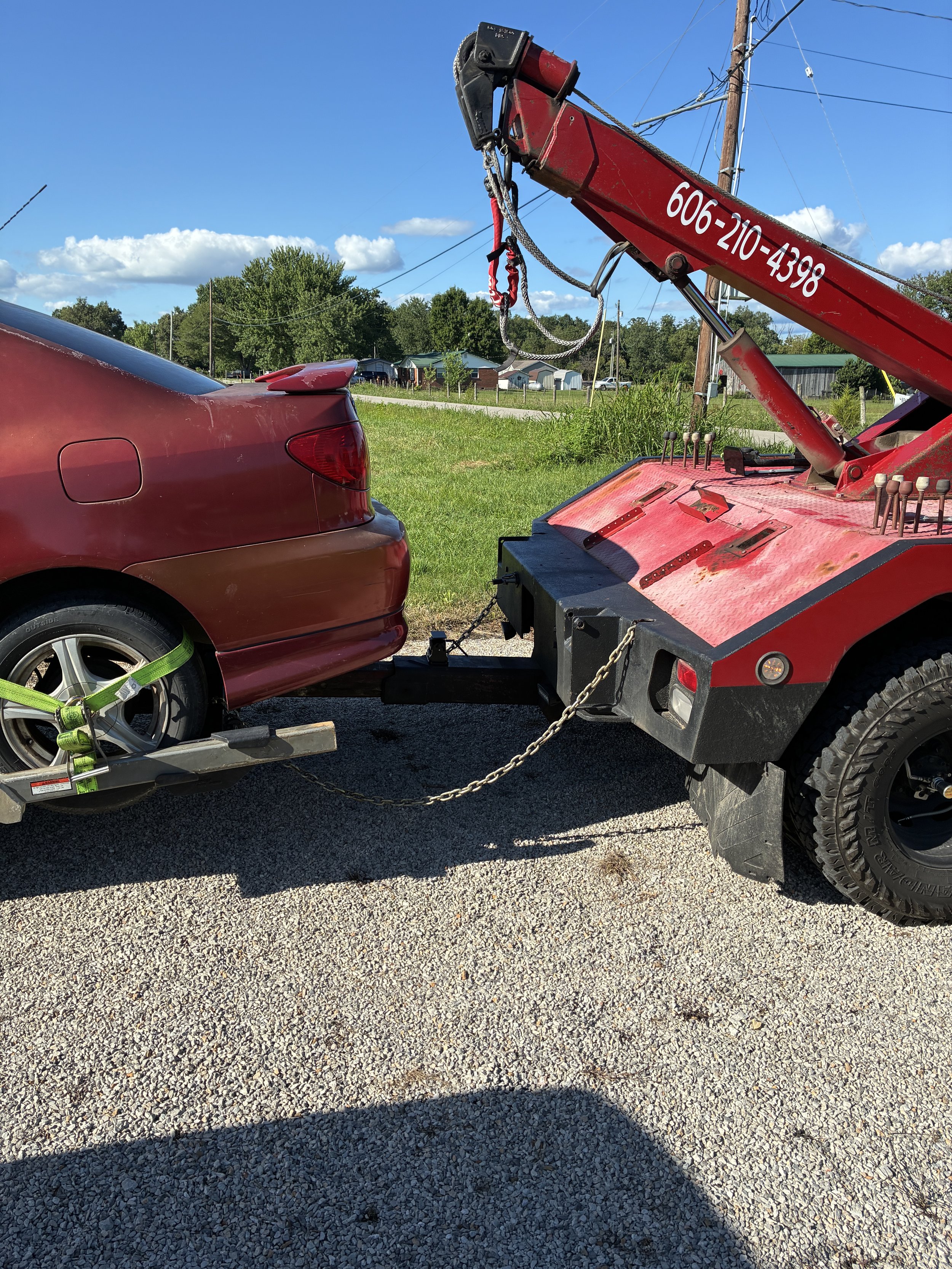 A red tow truck with a red Toyota Corolla attached to it. The pickup has a green strap securing it to the trailer. The background shows a rural neighborhood with houses, trees, and utility poles under a blue sky with clouds.