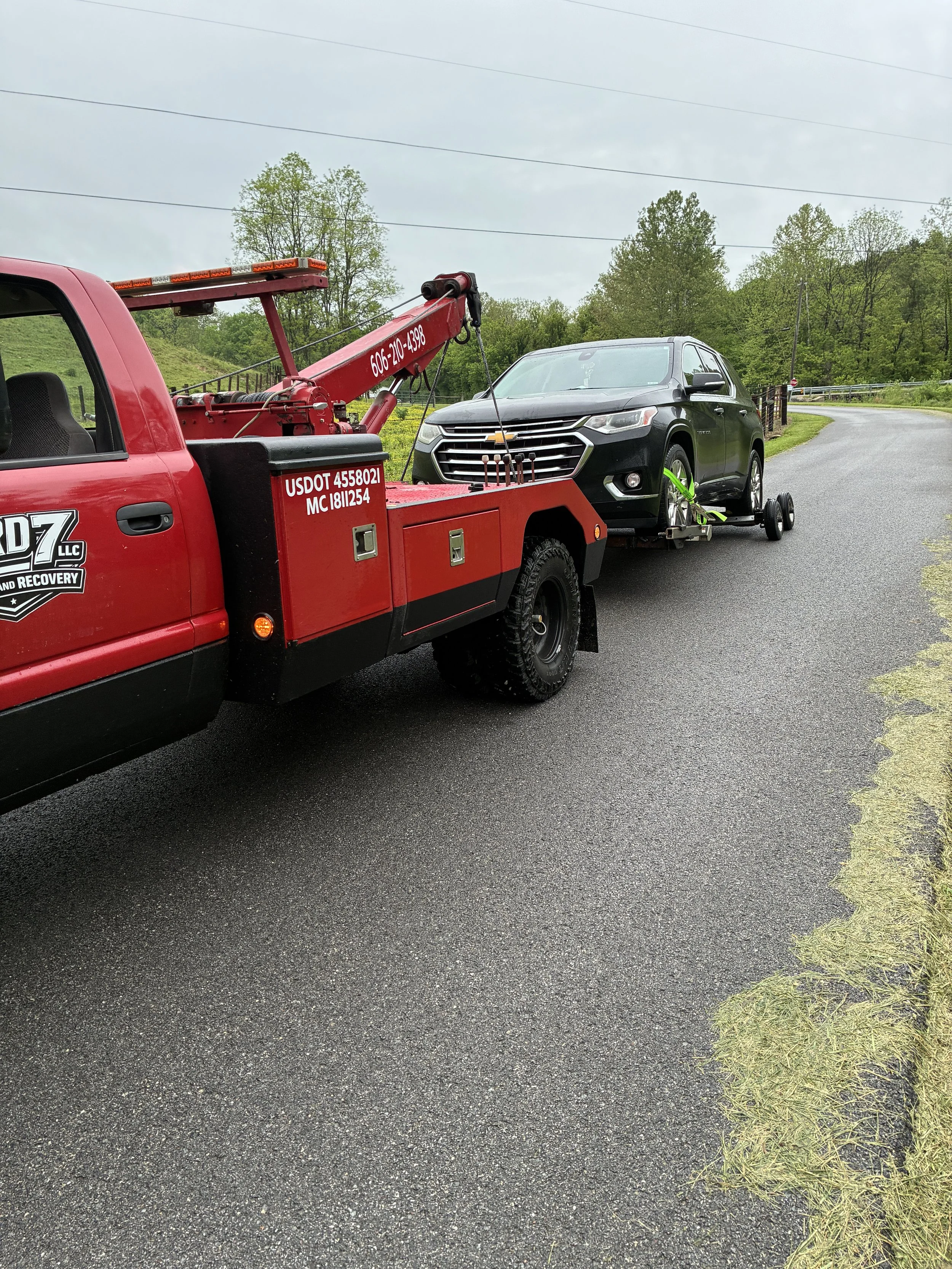 A red tow truck with a phone number and USDOT number is towing a black Chevrolet SUV on a curved paved road, with green trees and hills in the background and overcast sky.