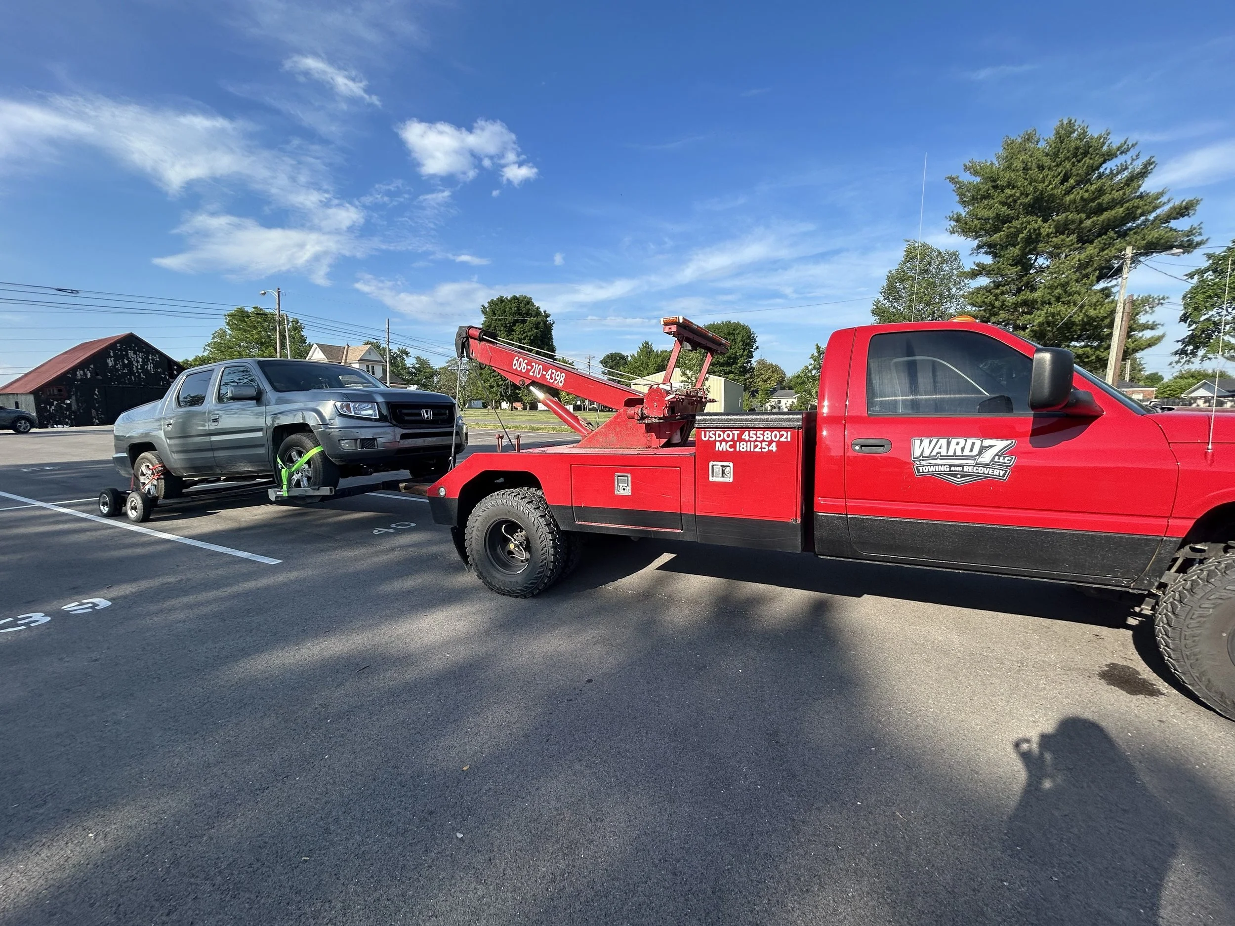 A red tow truck hauling a silver Honda Ridgeline truck in a parking lot with trees, buildings, and a clear blue sky in the background.