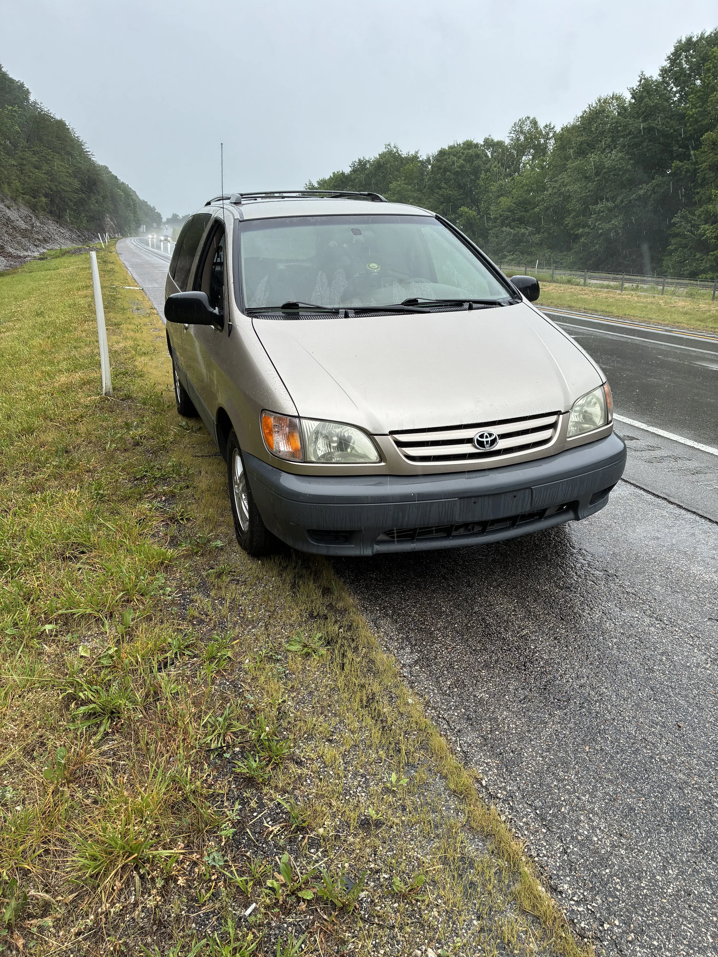 Silver minivan parked on the side of a wet interstate next to grass and guardrails during a rainy day.