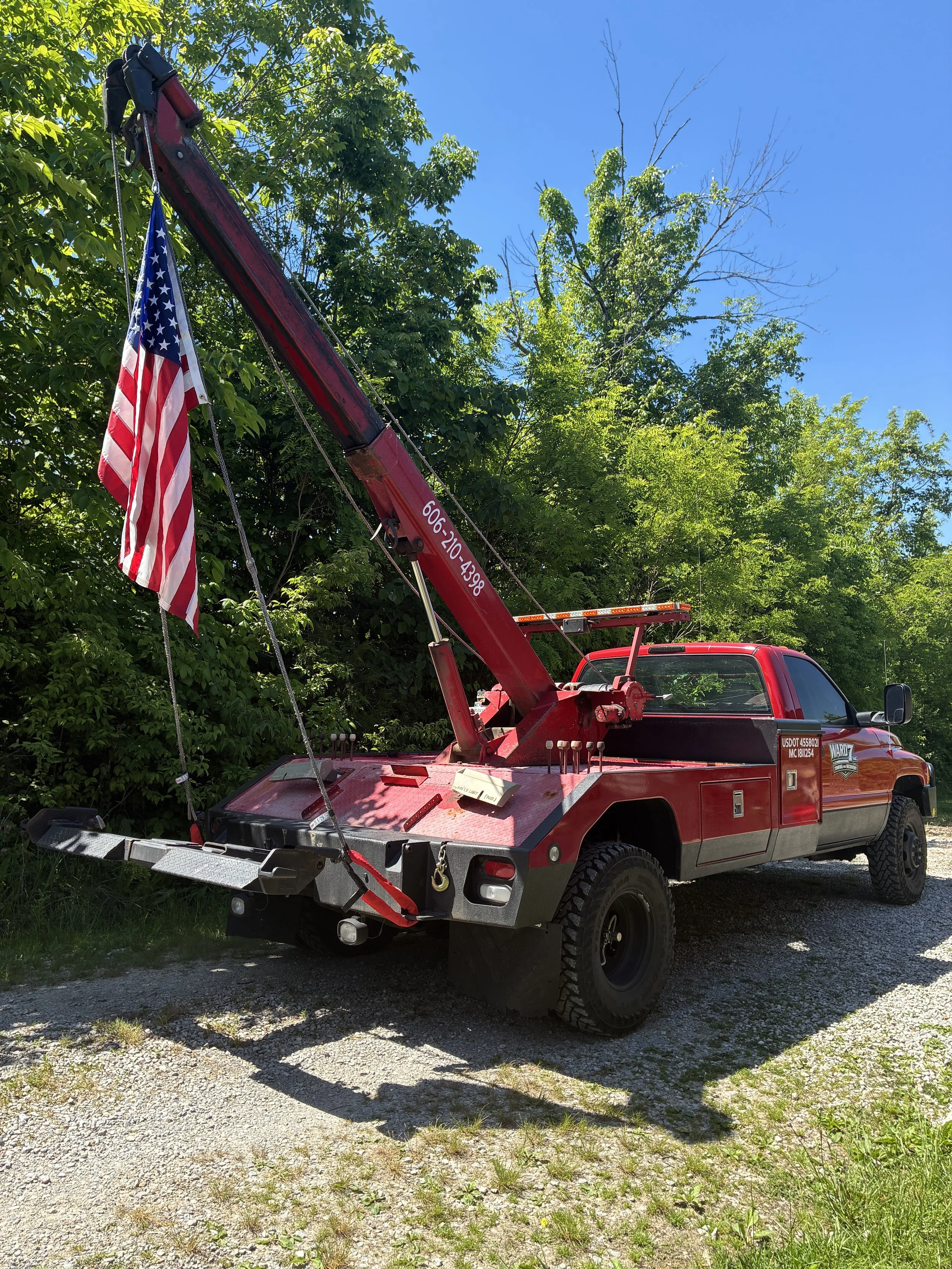 Red truck fitted with a crane, displaying an American flag, parked on a gravel surface with trees and a clear blue sky in the background.