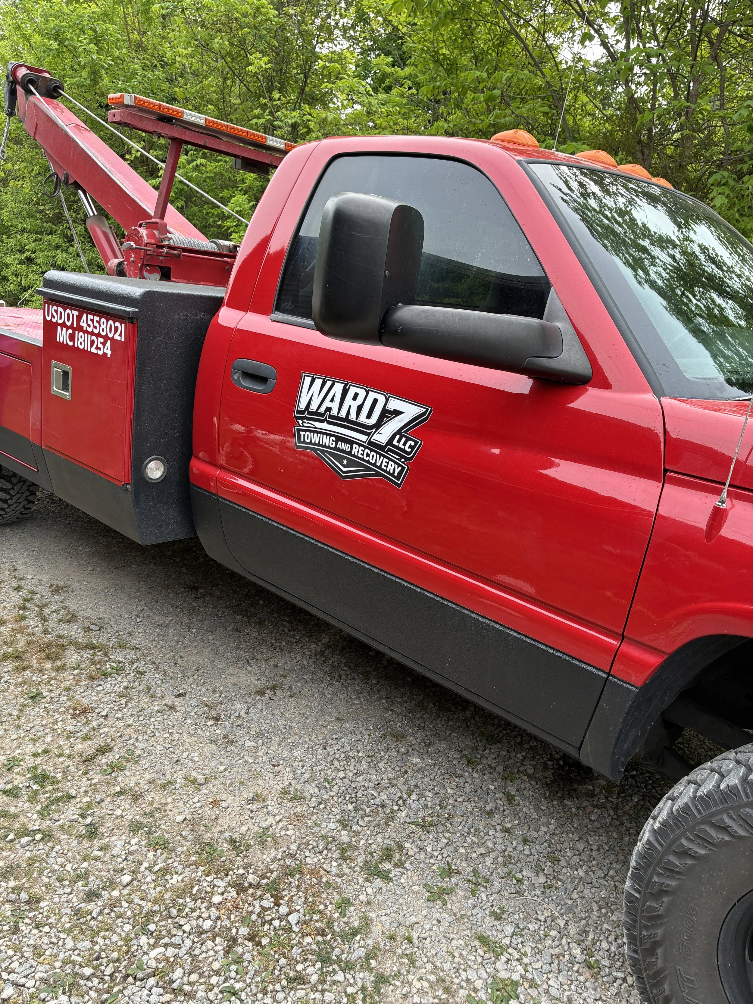 Red tow truck parked on a gravel surface with green trees in the background, displaying company branding and DOT information.