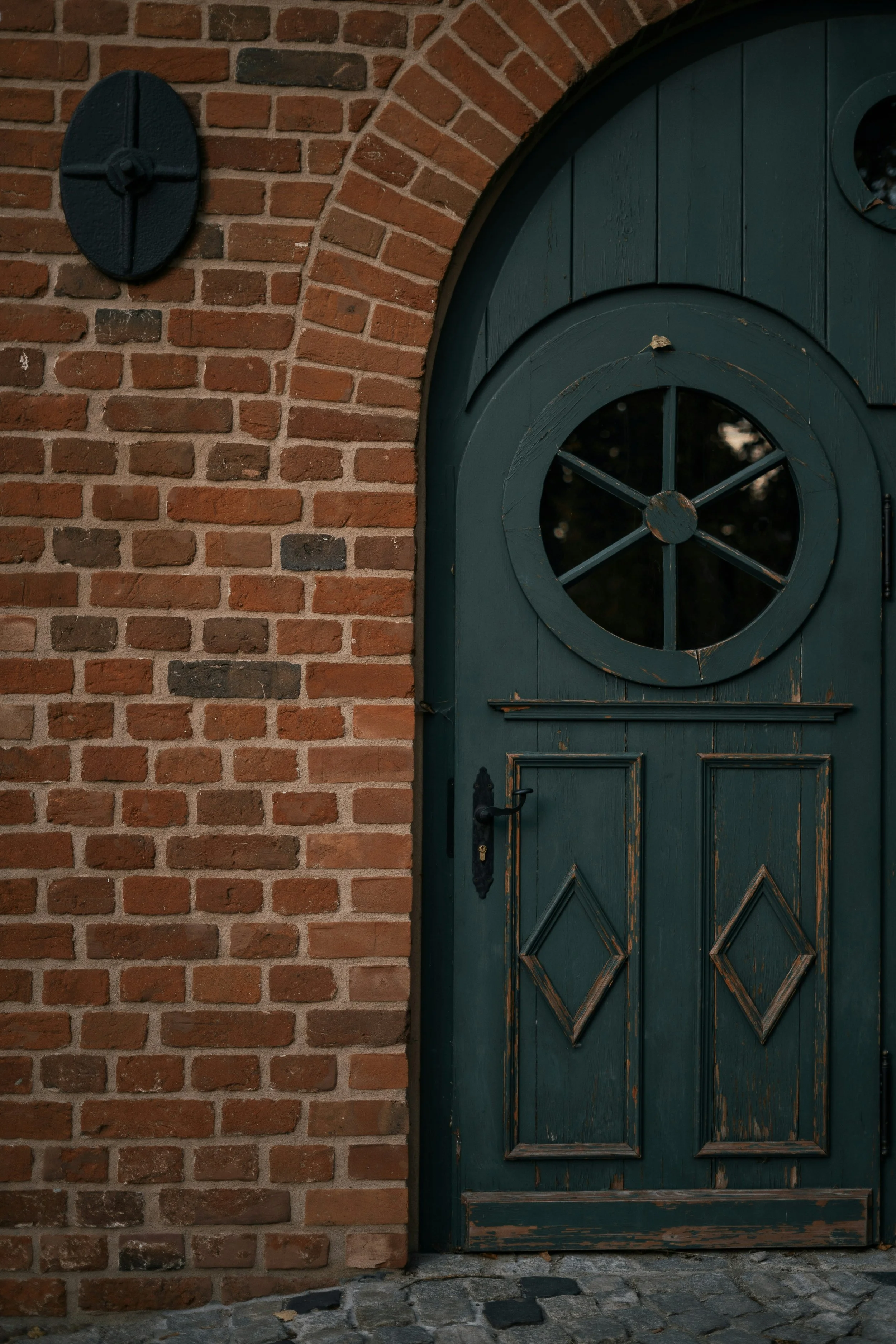 A rustic green wooden door with decorative carvings and a circular window, set in a red brick wall with an arched top, on a cobblestone ground.