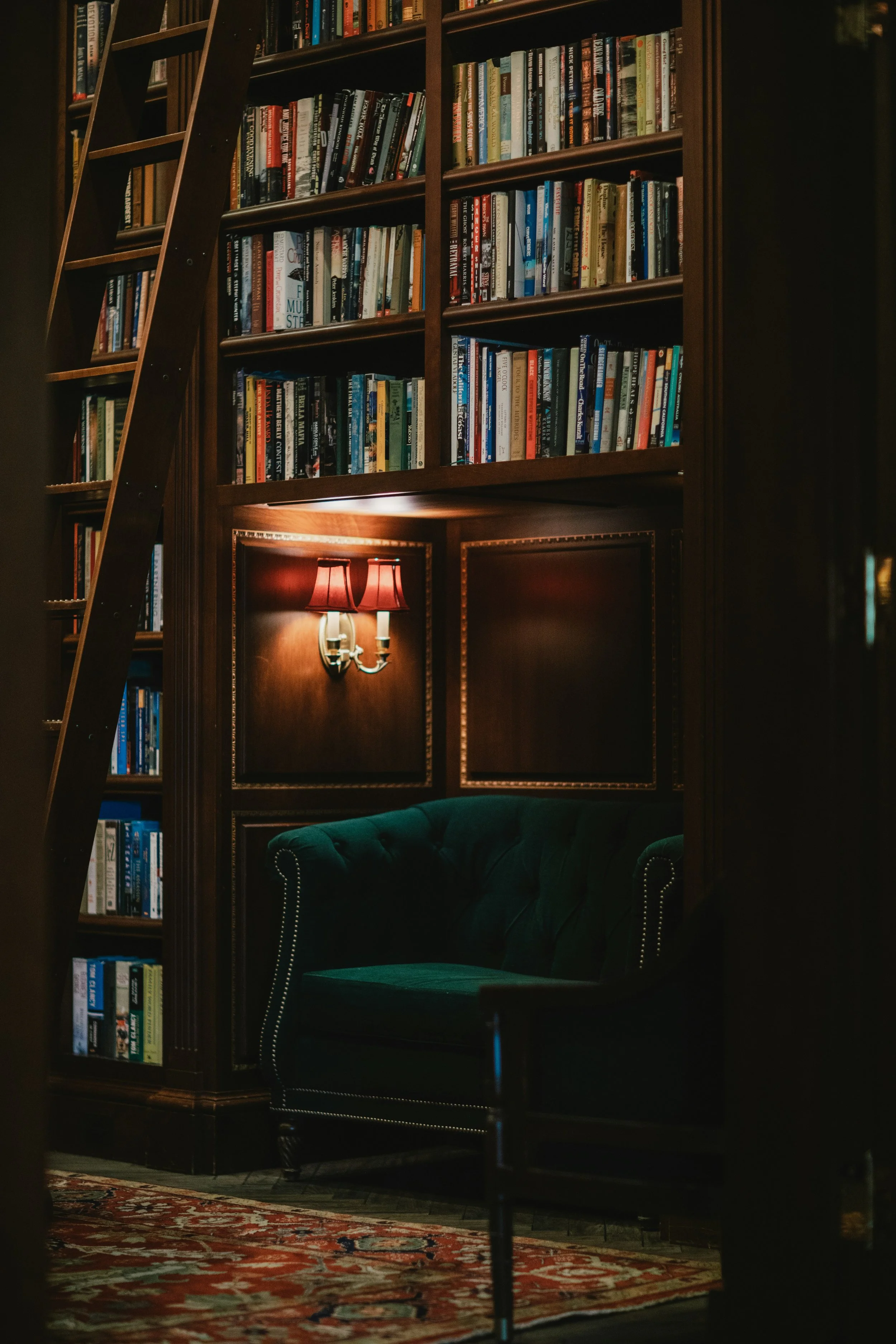 A dimly lit library or study with wooden bookshelves filled with books, a vintage red lampshade, and a green velvet armchair on a patterned rug.