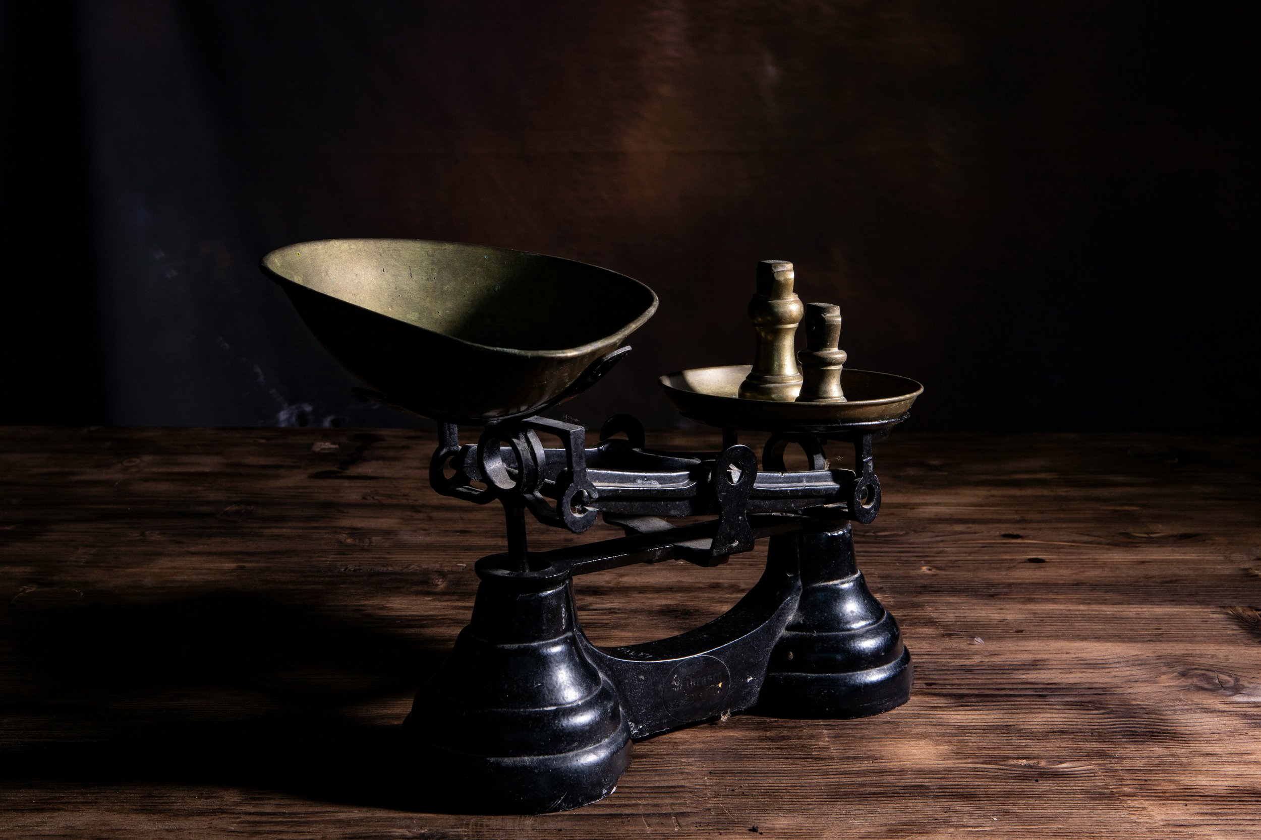An antique black weighing scale with brass weights on a metal dish, placed on a wooden surface with a dark background.