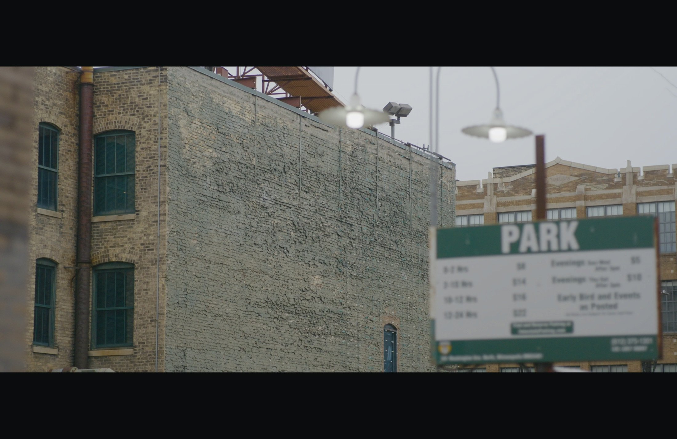 A city scene featuring an old brick building with tall, arched windows and a fire escape, metal street lighting, and a blurred parking sign in the foreground.