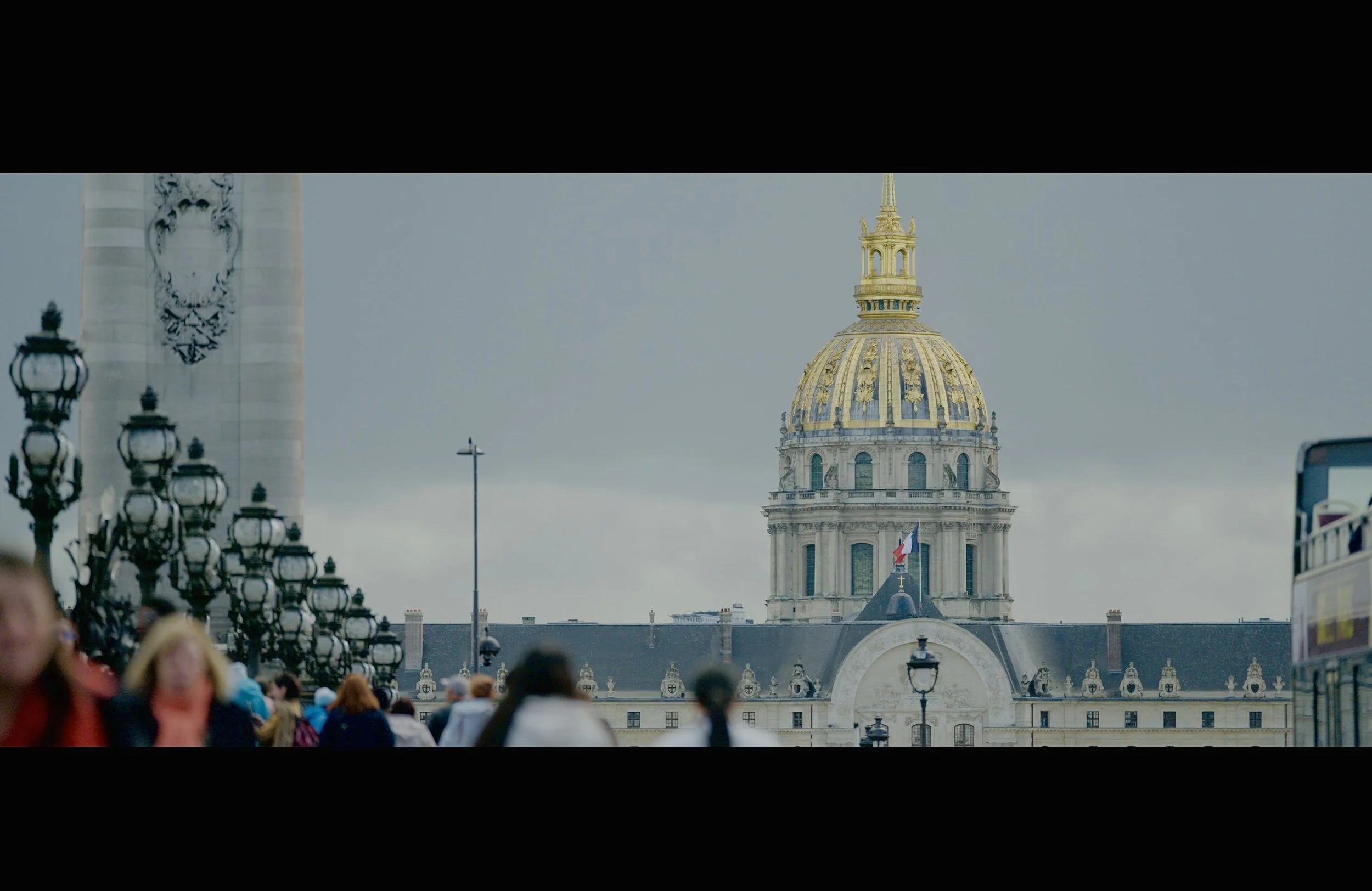 A crowded city street with ornate street lamps, leading to a historic building with a golden-domed roof and a French flag, under a cloudy sky.