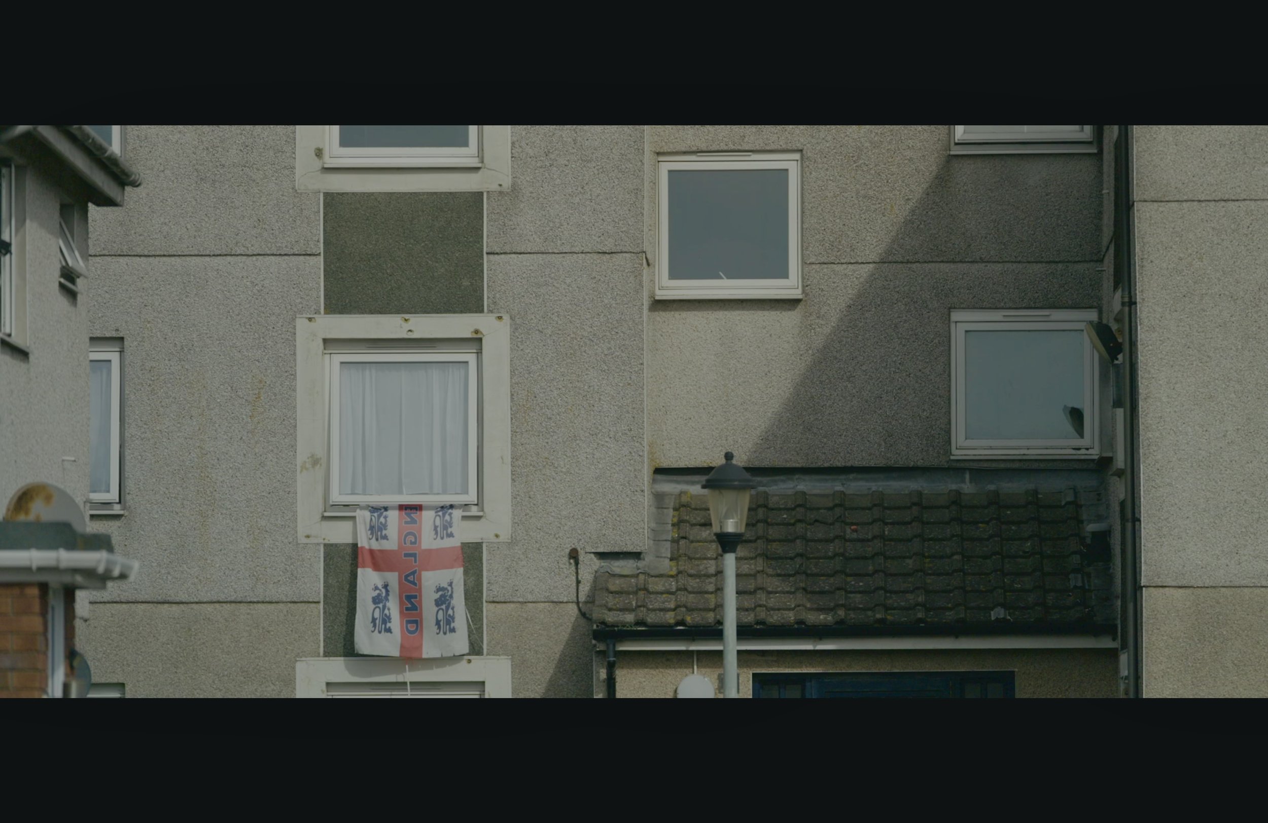 Close-up of the exterior wall of a residential building with multiple windows, a lamp post, and a flag hanging from a balcony, on a partly cloudy day.