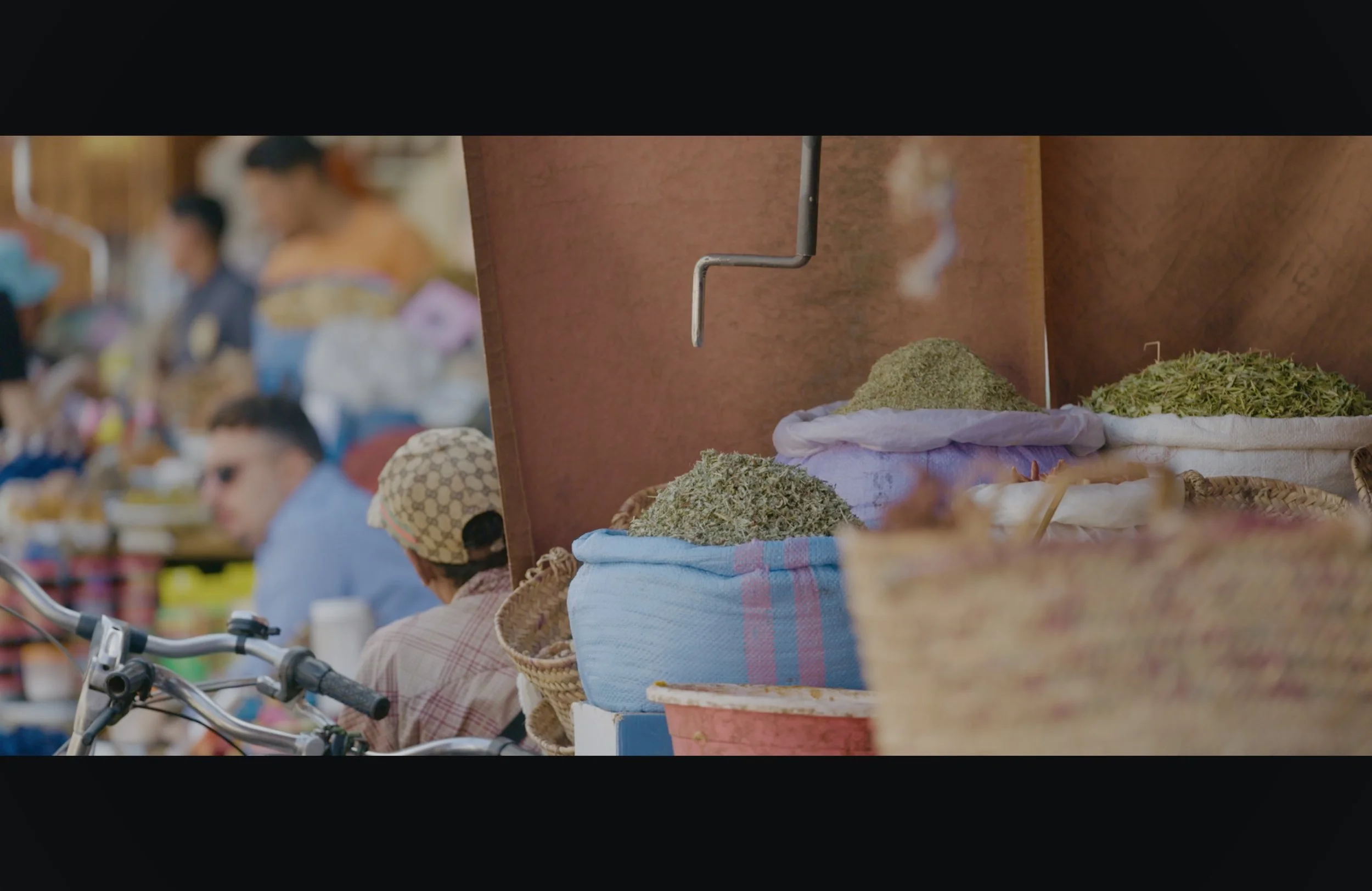Market scene with people, baskets of dried herbs or plants, and vendors in the background.