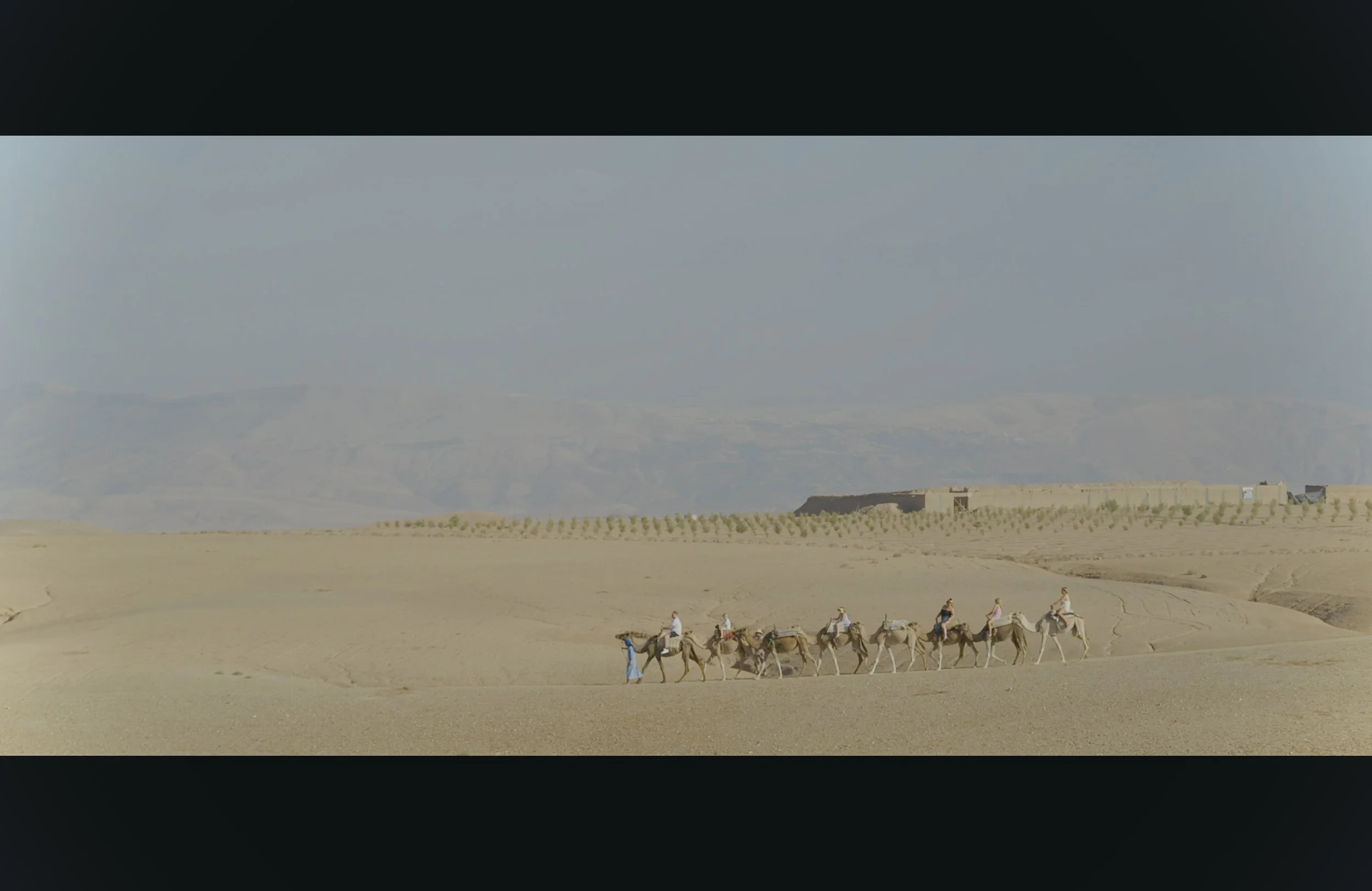 A group of people riding camels in a desert landscape, with mountains in the background and a building on the right side.