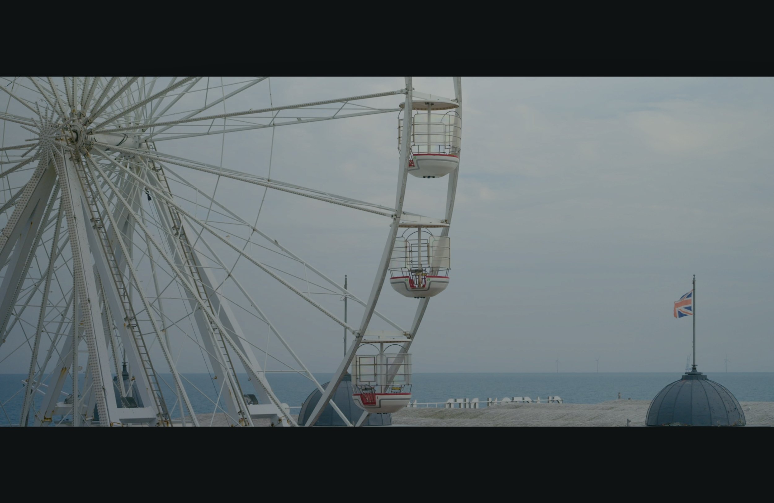 A Ferris wheel with red and white passenger cabins near the ocean, with a cloudy sky and a Union Jack flag flying in the background.