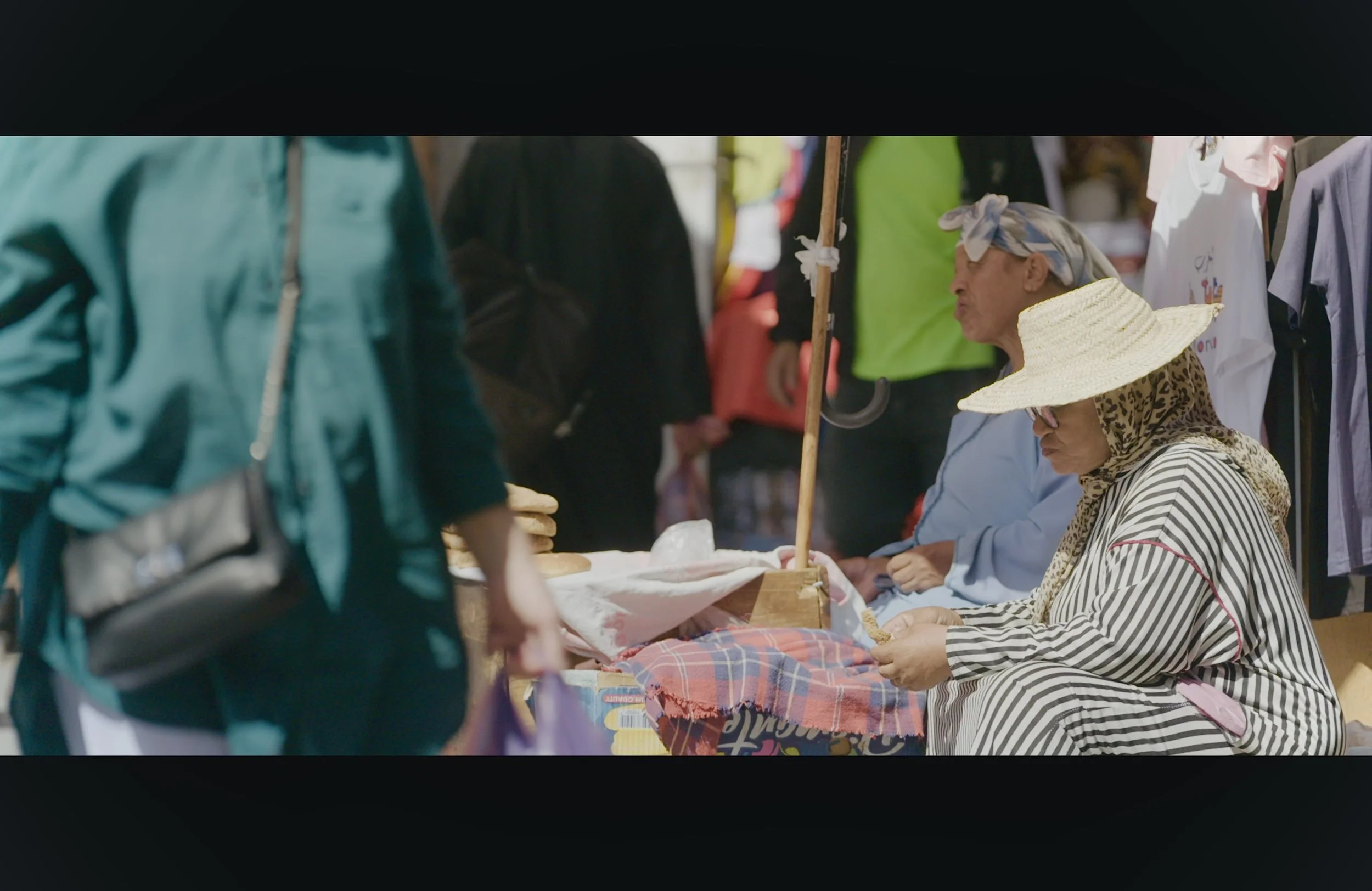 A woman with a striped shirt and animal print scarf and a sun hat sitting at a market stall, talking to a person in a green shirt, with other market stalls and shoppers visible in the background.