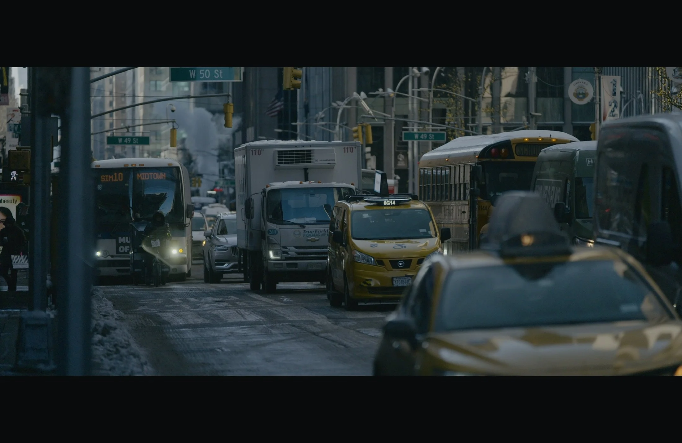 City street scene with multiple vehicles including buses and taxis titled 'W 49 St' and 'W 50 St', with buildings, traffic lights, and signs visible.