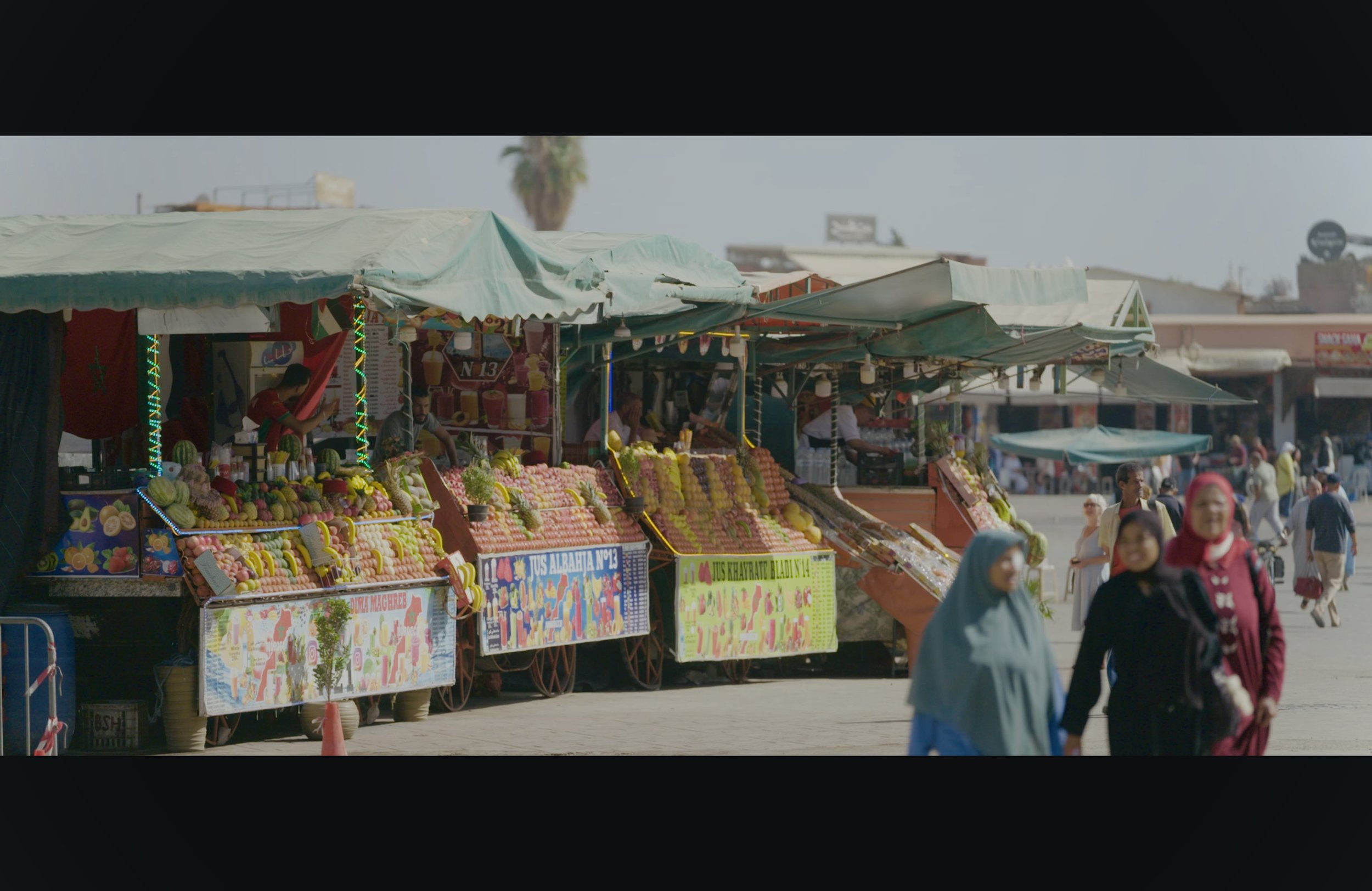 Street market with stalls selling fruits and produce, with people walking and browsing, under umbrellas, in a busy outdoor area.