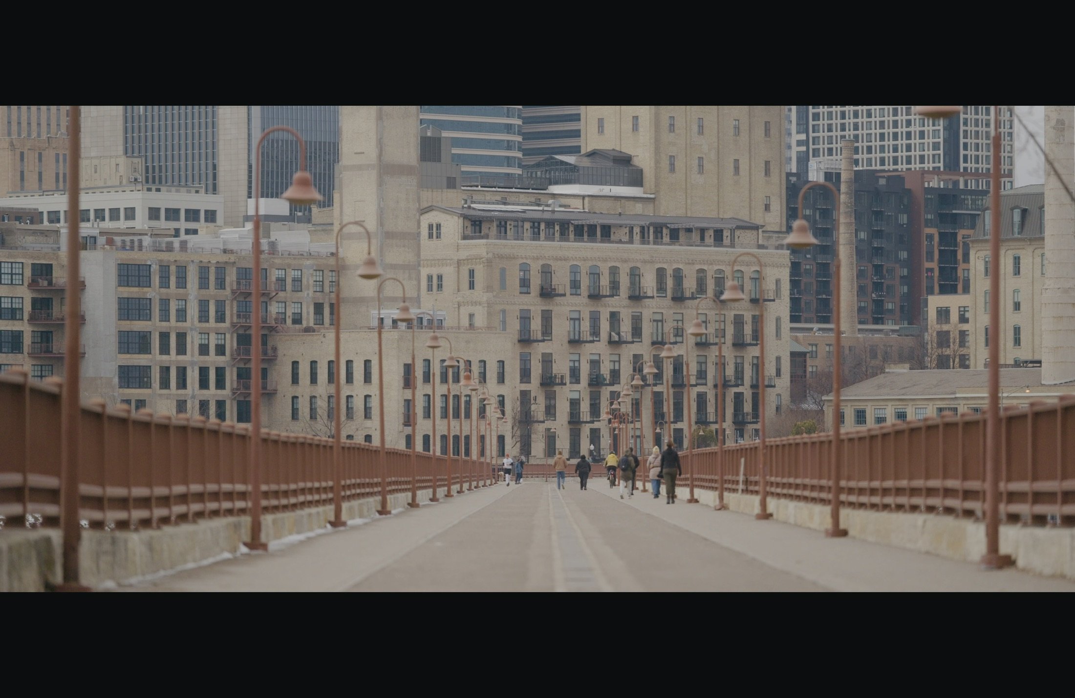 A cityscape view from a pedestrian bridge with people walking, overlooking multiple old and modern buildings, lampposts along the bridge, and tall skyscrapers in the background.