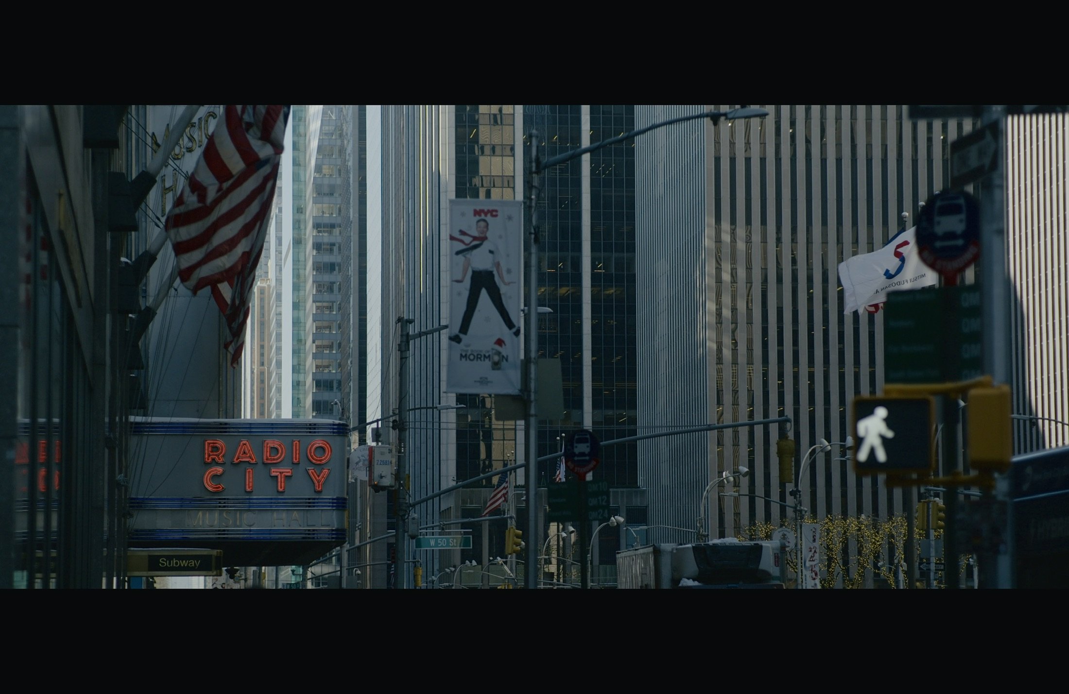 City street scene with tall buildings, American flags, and advertising signs including 'Radio City' and a Manhattan billboard. Pedestrian crossing sign and traffic lights are visible.