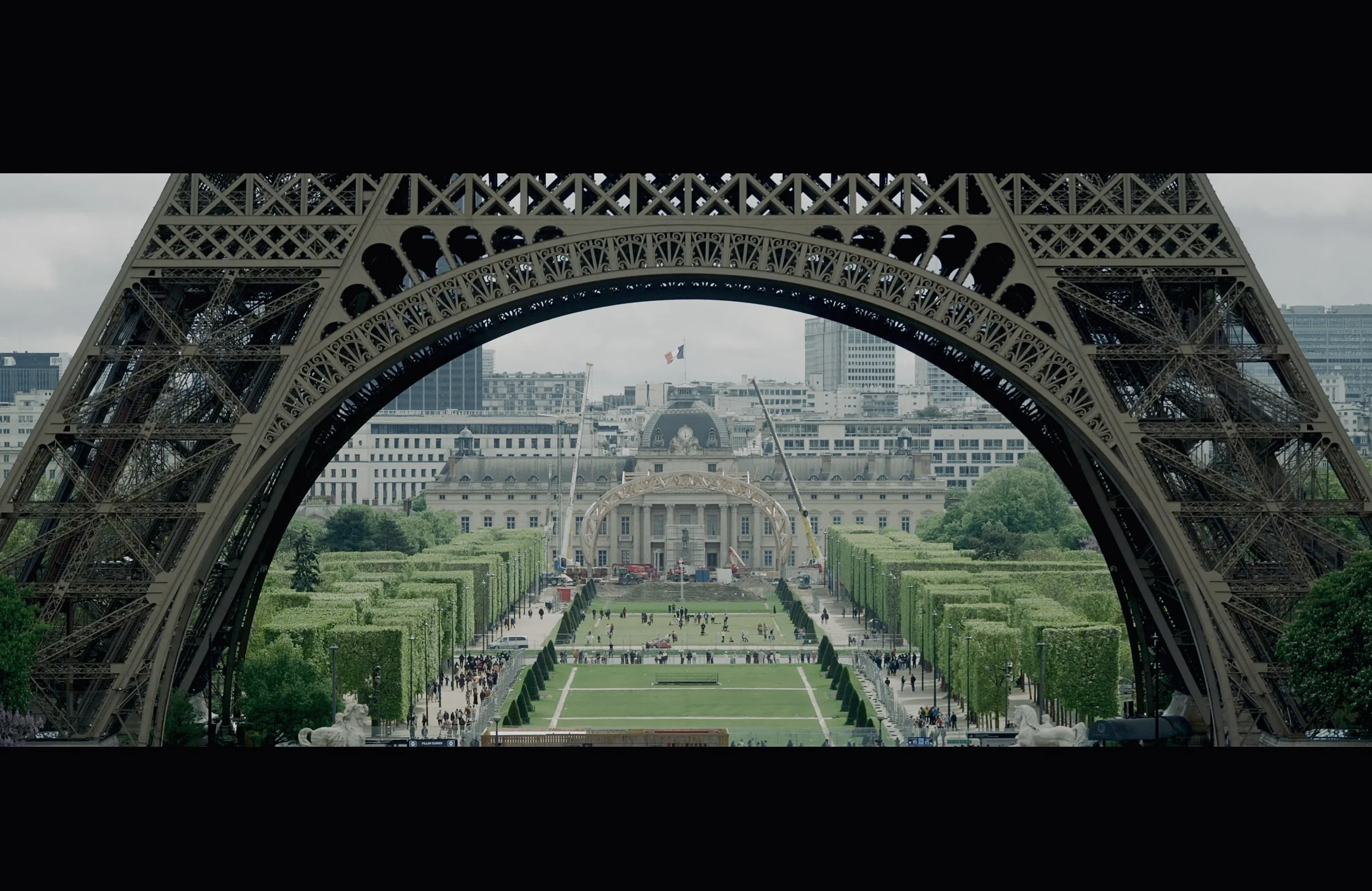 The Eiffel Tower viewed from its base, with the city of Paris in the background, including buildings and the French flag. The foreground shows a green garden area with people walking and sitting.