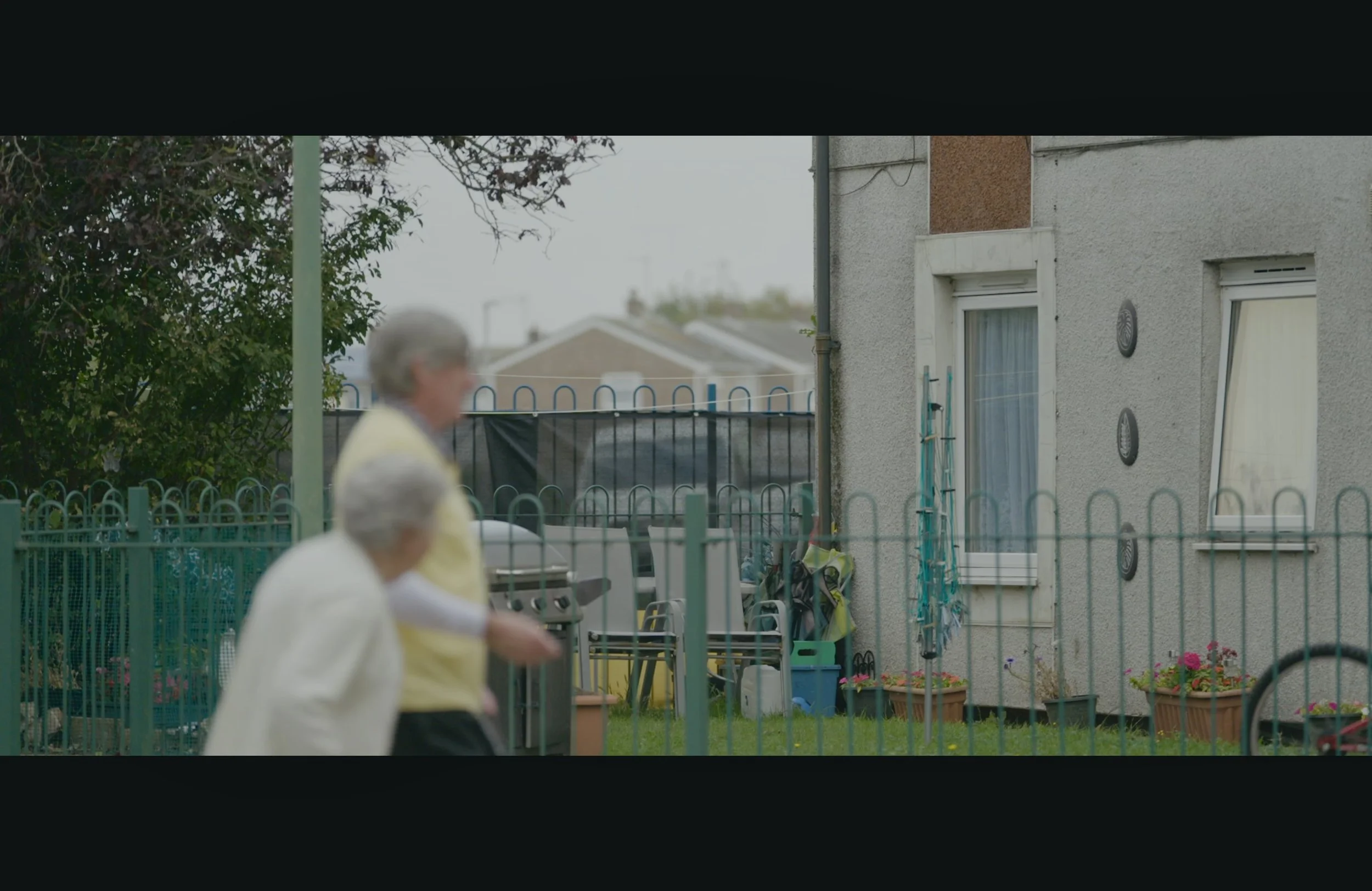 Two elderly women walking past a residential backyard with a mint-green fence, potted plants, and house windows in the background.