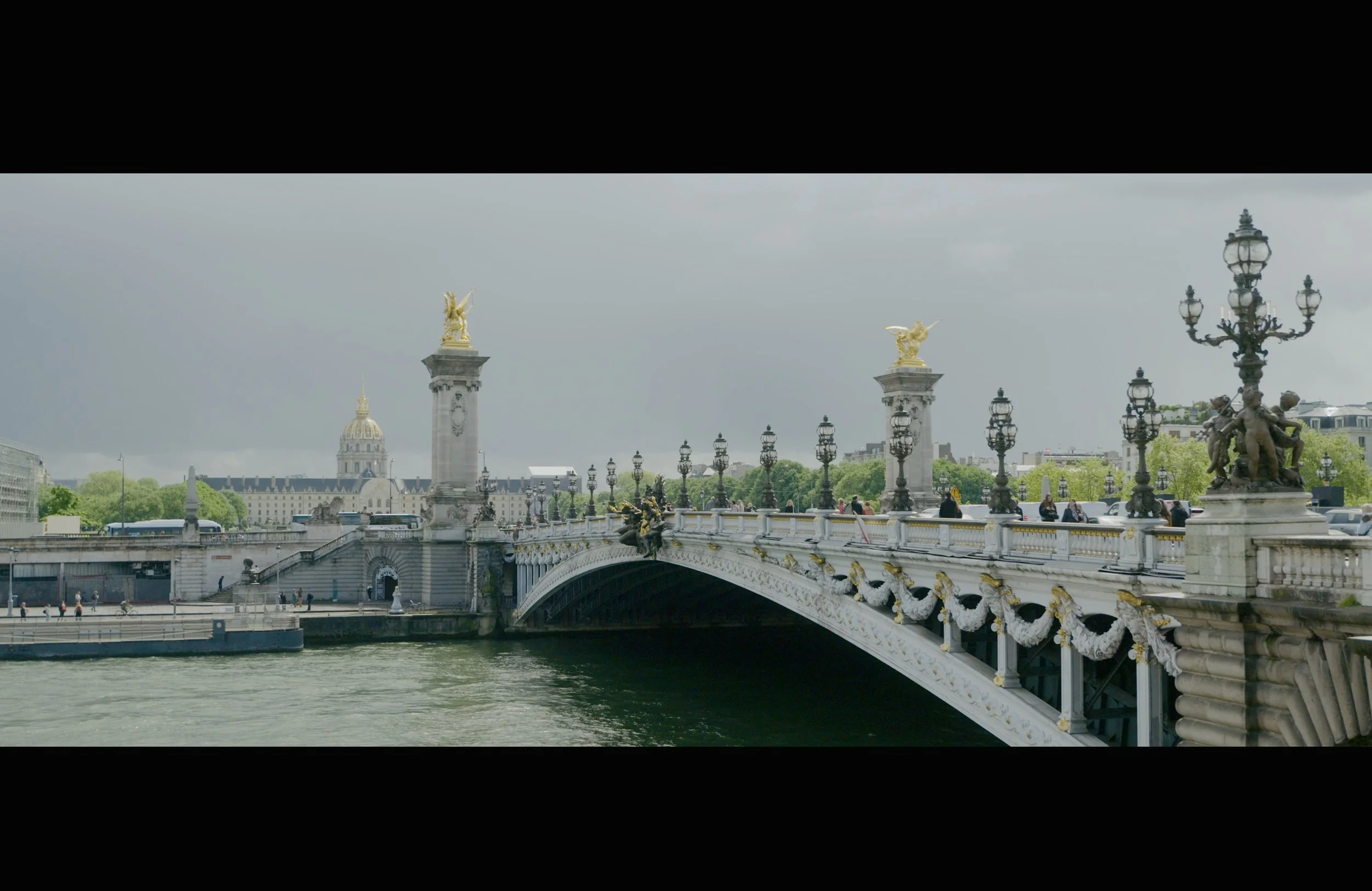 Paris bridge over the Seine River with ornate lamp posts, sculptures, and gilded statues, and historic buildings in the background under a cloudy sky.