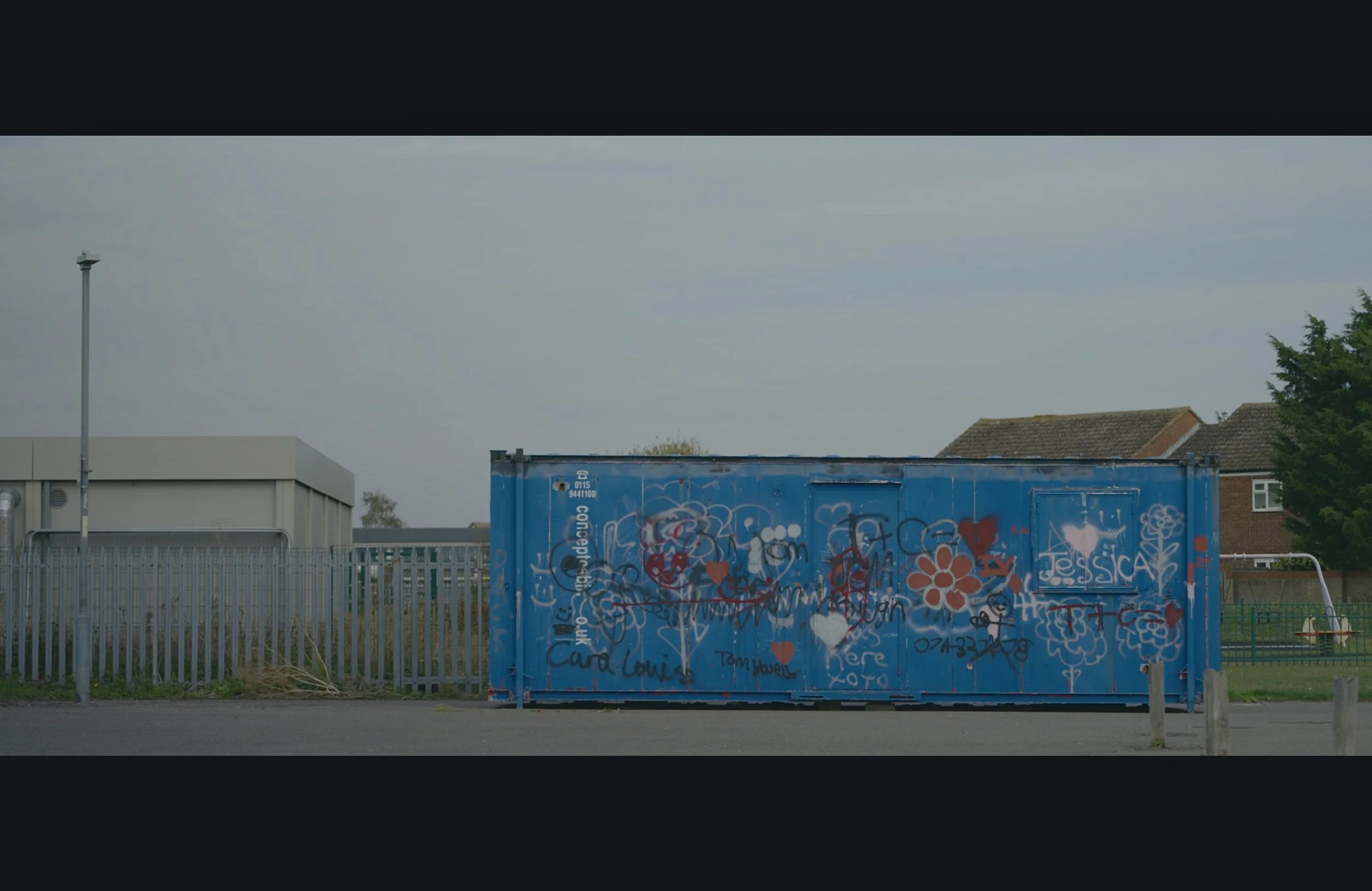 A blue graffiti-covered storage container in front of a metal fence, with residential houses and a tree in the background under a cloudy sky.