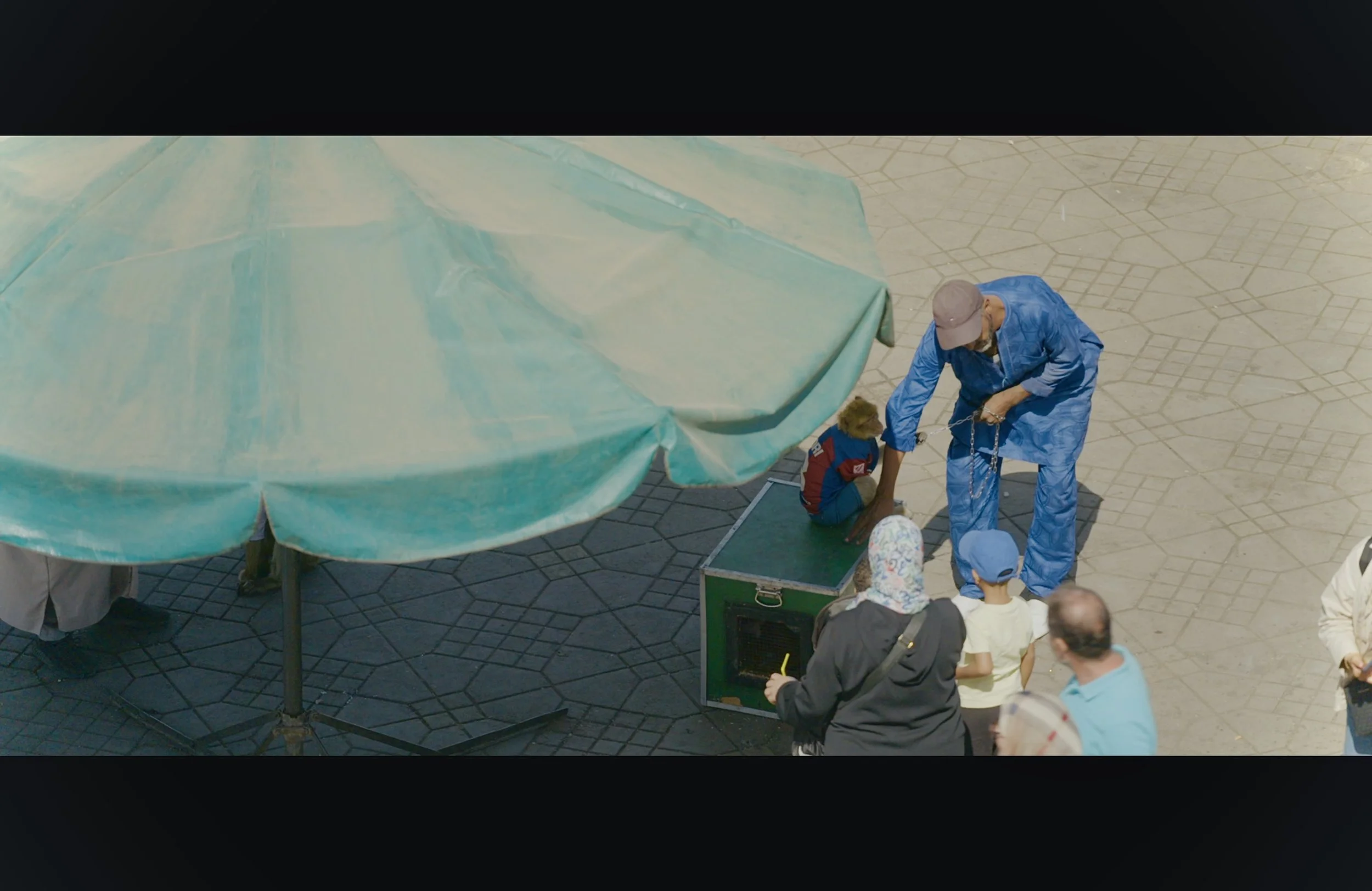 A person dressed in blue standing on the street with a chain in their hand, surrounded by a small group of people, some of whom are children, gathered around a small black box or speaker. A green umbrella and a light-colored tiled pavement are also v